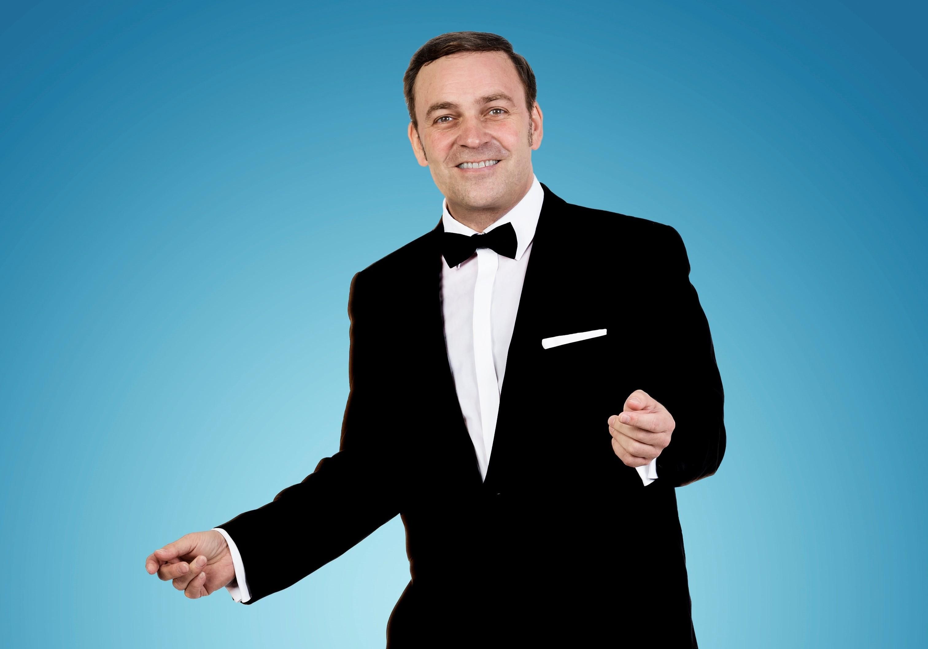 Martin Schmid in schwarzem Smoking mit schwarzer Fliege am Hals und weißem Hemd in legerer, swingender Tanzpose vor türkisfarbenem Hintergrund mit Lichtspot in Szene gesetzt.