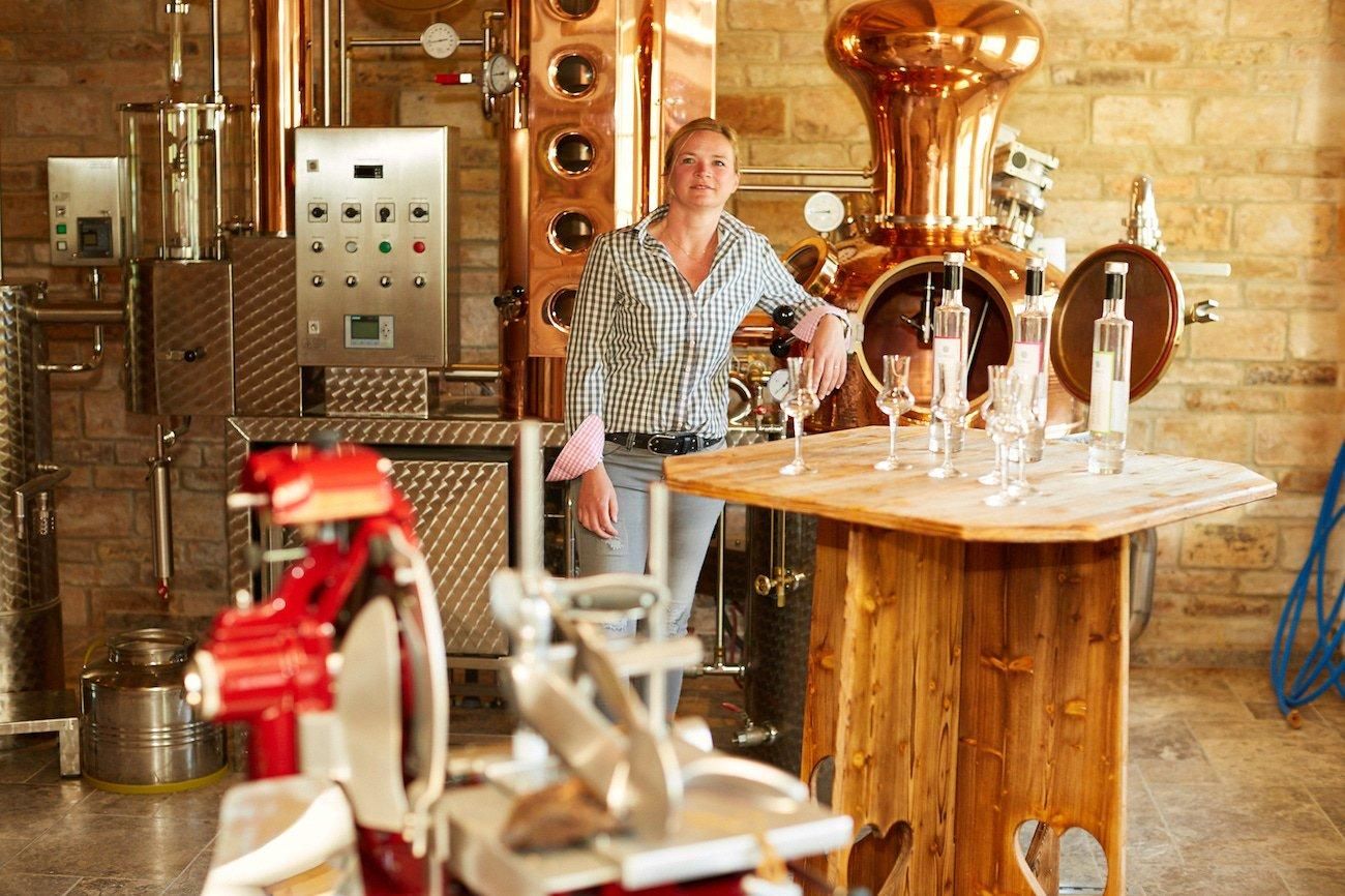 Eine Frau steht lächelnd in einer Brennerei, umgeben von glänzenden Kupferkesseln und Destillationsgeräten.