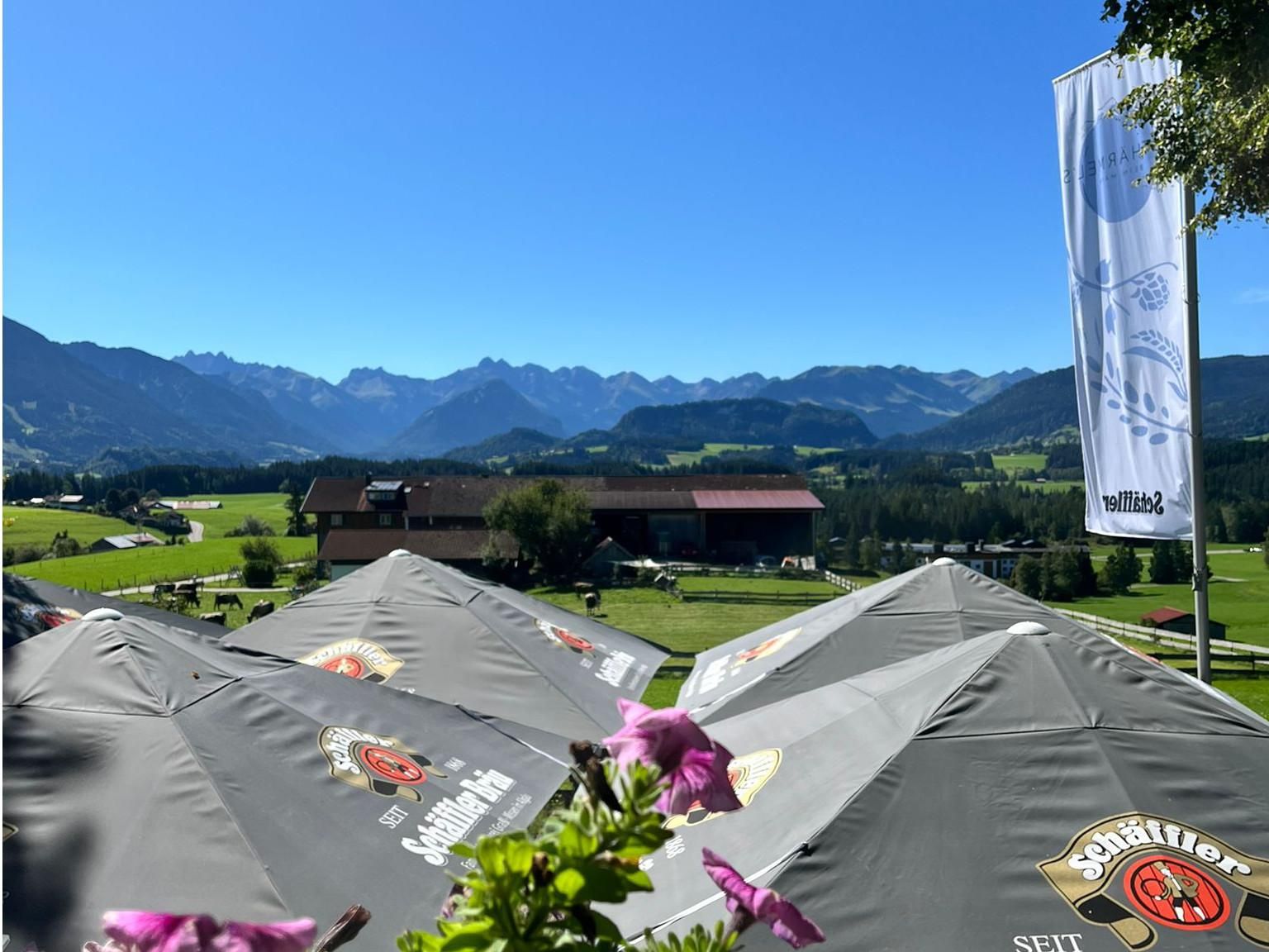 Blick auf Terrasse mit Sonnenschirmen des Bauerngasthofes Rothärmel's beim Mayr in Ofterschwang, im Hintergrund Bergpanorama und grüne Wiesen.