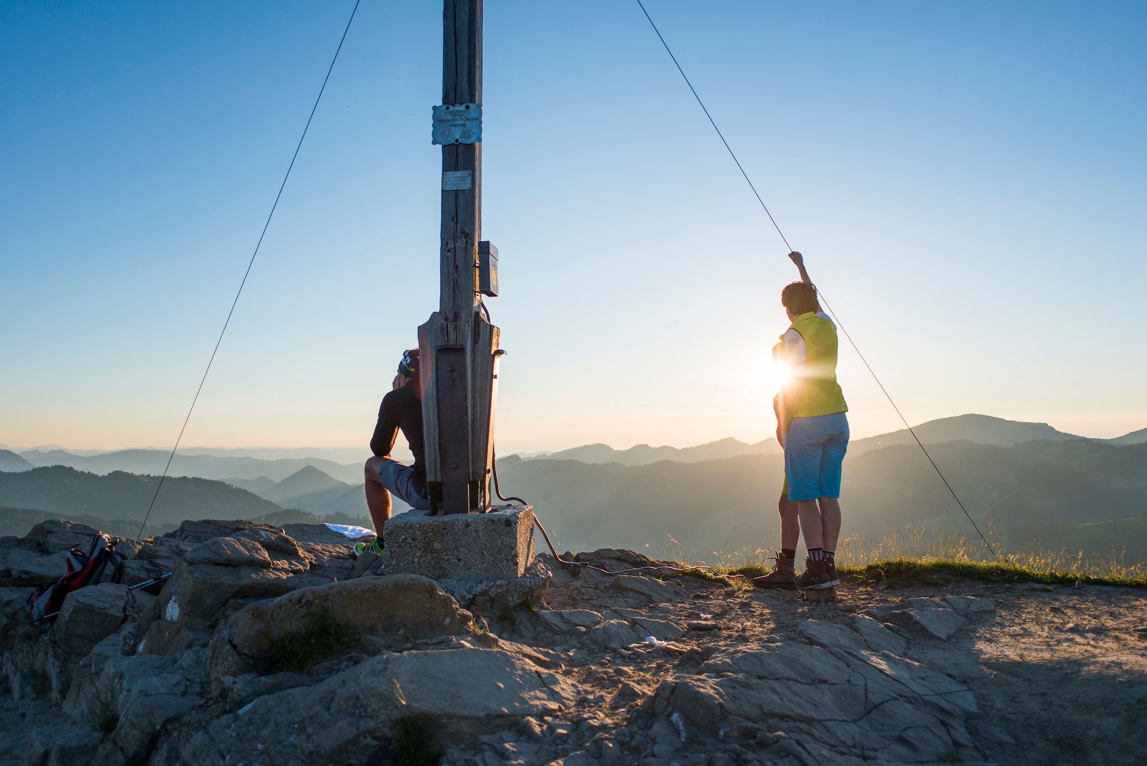 Zwei Wanderer genießen Sonnenaufgang am Gipfelkreuz Riedberger Horn