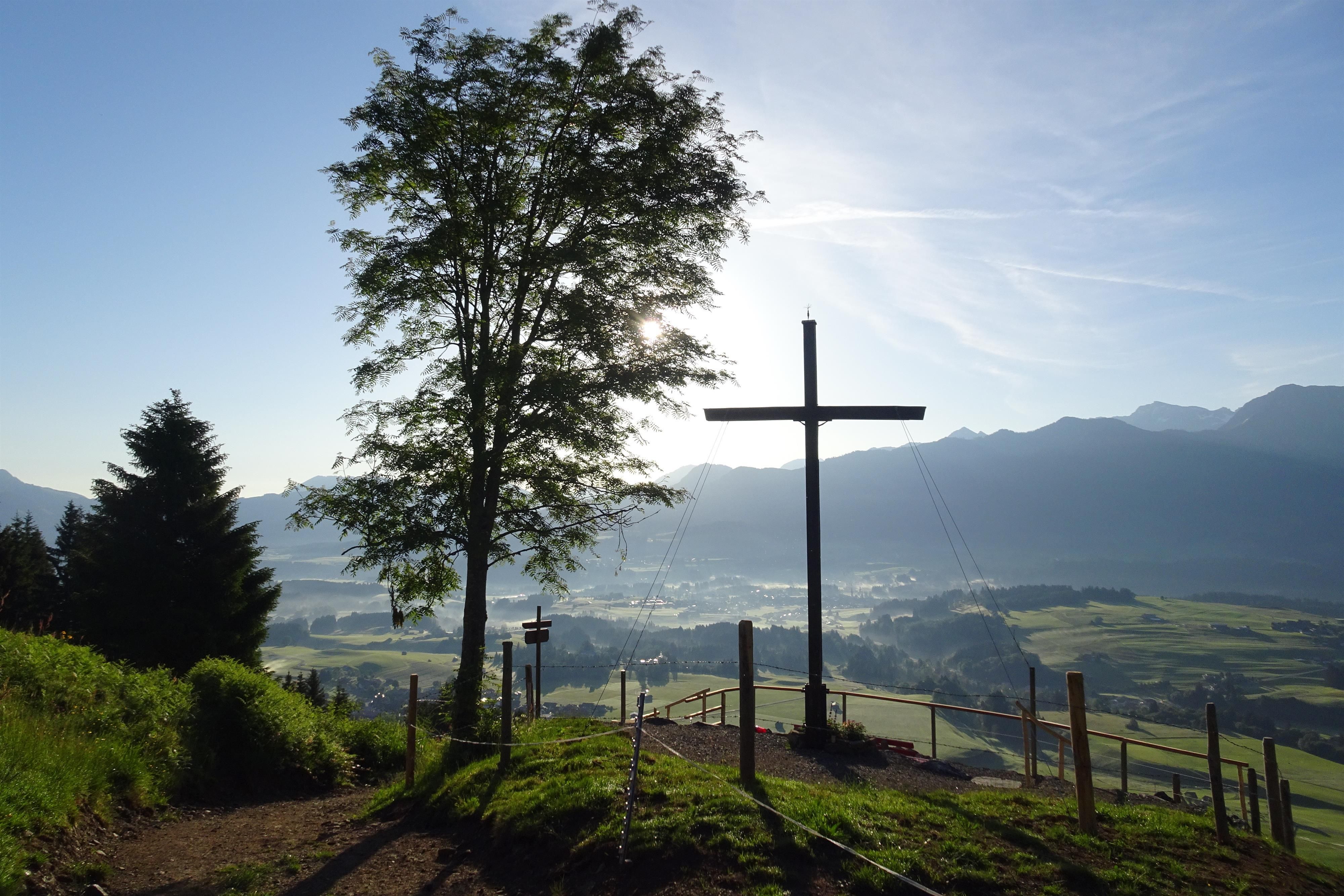 Ein großes, dunkles Holzkreuz auf einem Berg. Im Hintergrund sind bewaldete Hügel und eine weite, nebelverhangene Ebene mit Häusern zu sehen.