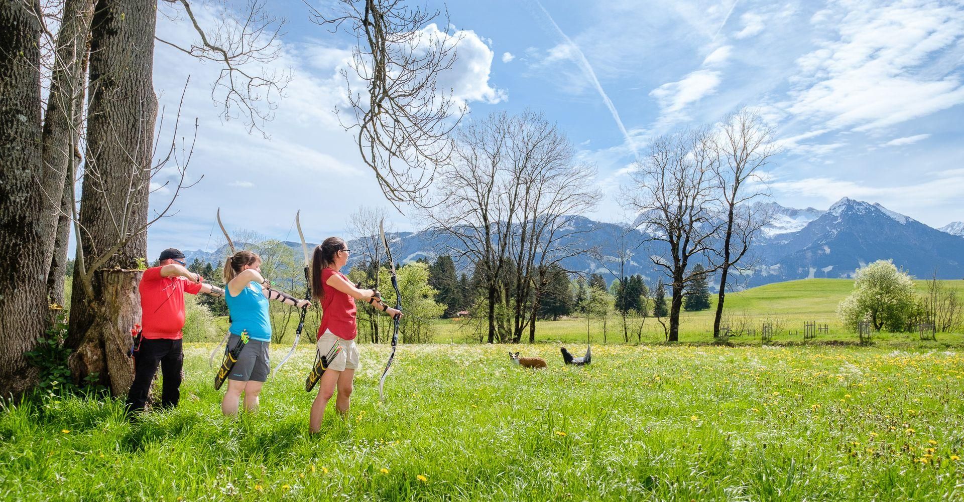 Drei Personen beim Bogenschießen auf einer Wiese, im Hintergrund Berge.