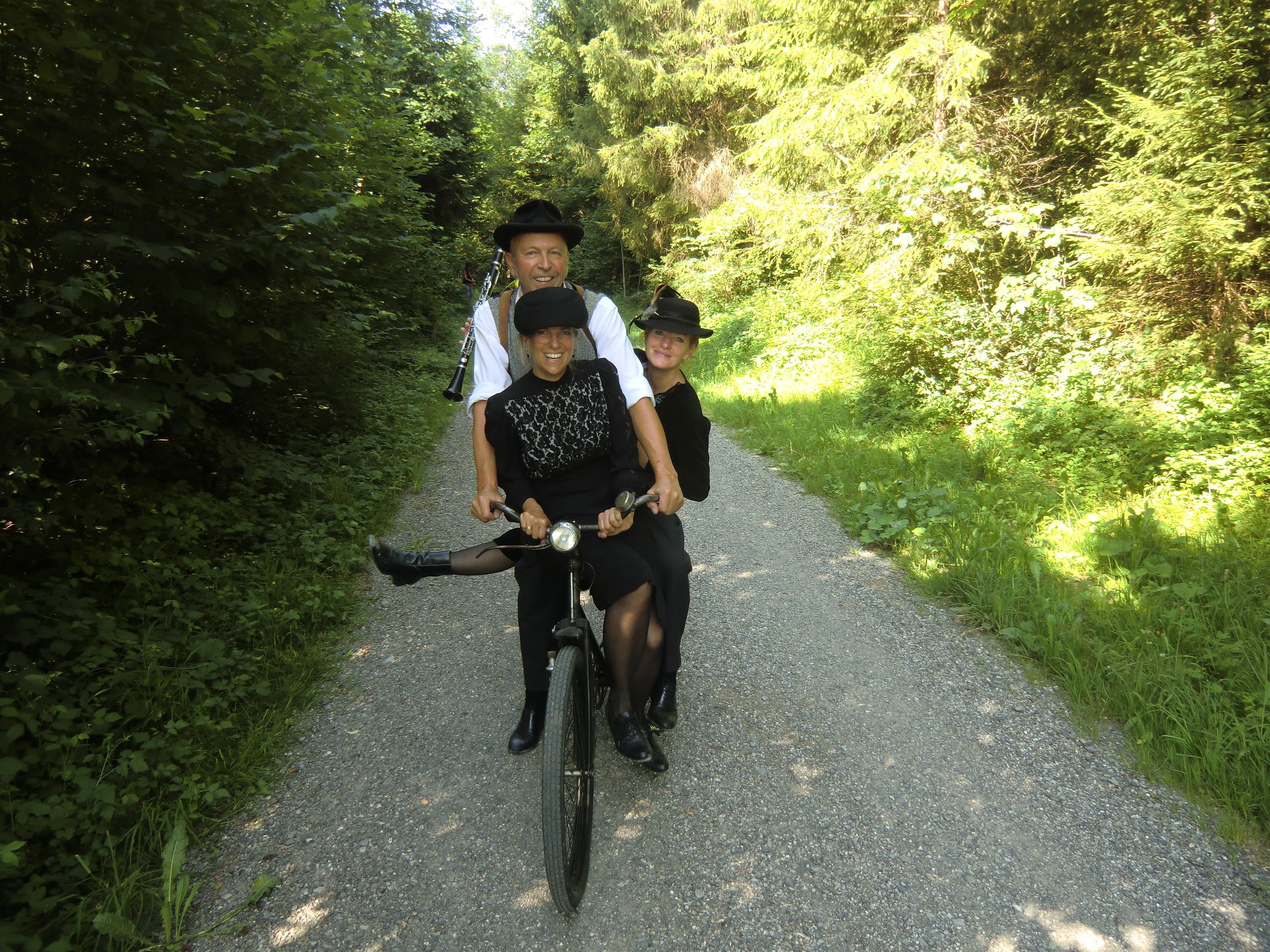 Ein Mann und zwei Frauen fahren auf einem Fahrrad einen Schotterweg entlang. Eingerahm von Wald. Die Personen tragen historische Kleidung.