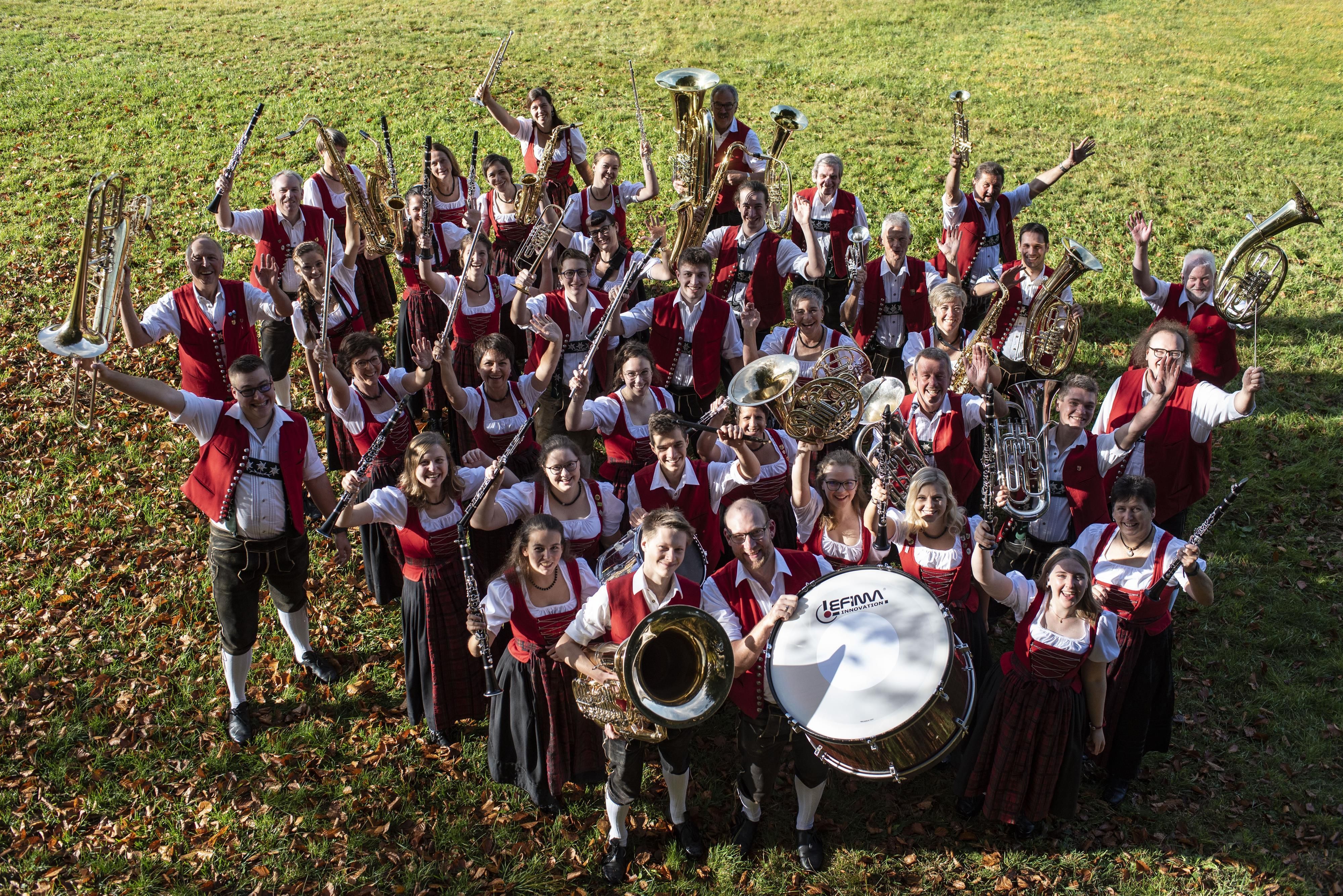 Foto von oben: die Blaskapelle Fischen auf einer Wiese, mit roten Westen oder Dirndloberteilen, die Insturmente dabei.