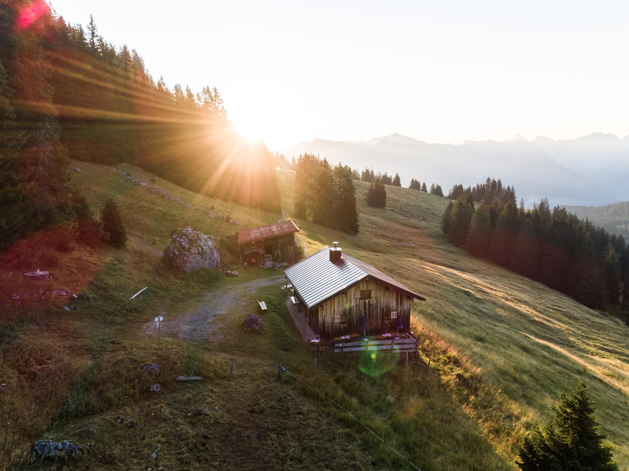 Obere Gund Alpe im Morgenlicht: Holzhütte am Hang, umgeben von grüner Wiese, Bäumen, Bergpanorama, Sonnenstrahlen.