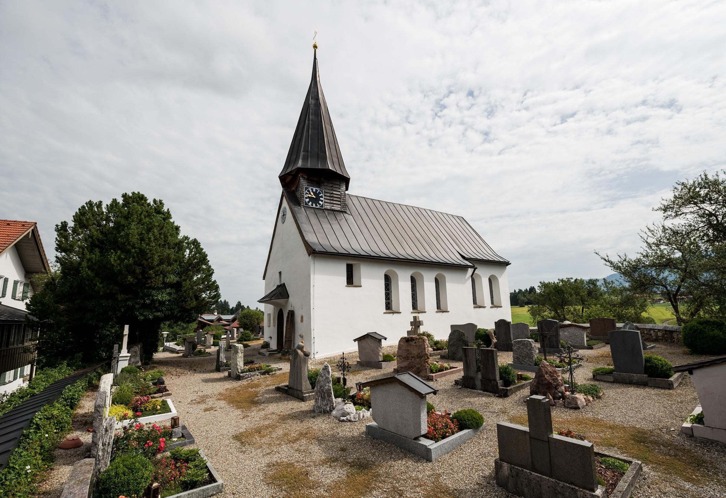 Weiße Kirche mit grauem Dach und spitzem Turm mit Uhr. Umgeben von einem Friedhof mit vielen Grabsteinen und Blumen.