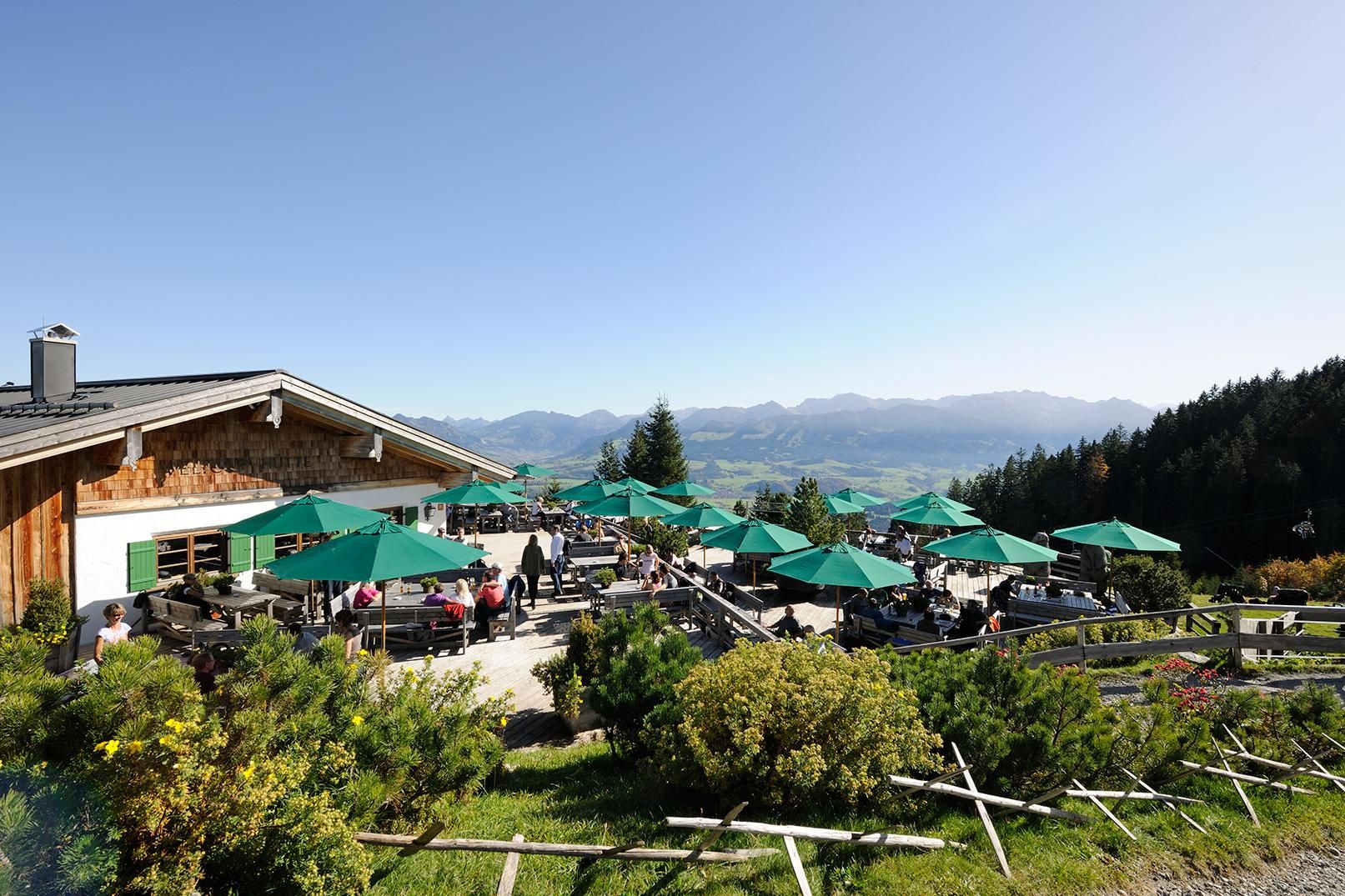 Weltcup Hütte Ofterschwang. Terasse mit grünen Sonnenschirmen und Menschen sitzen in der Sonne.