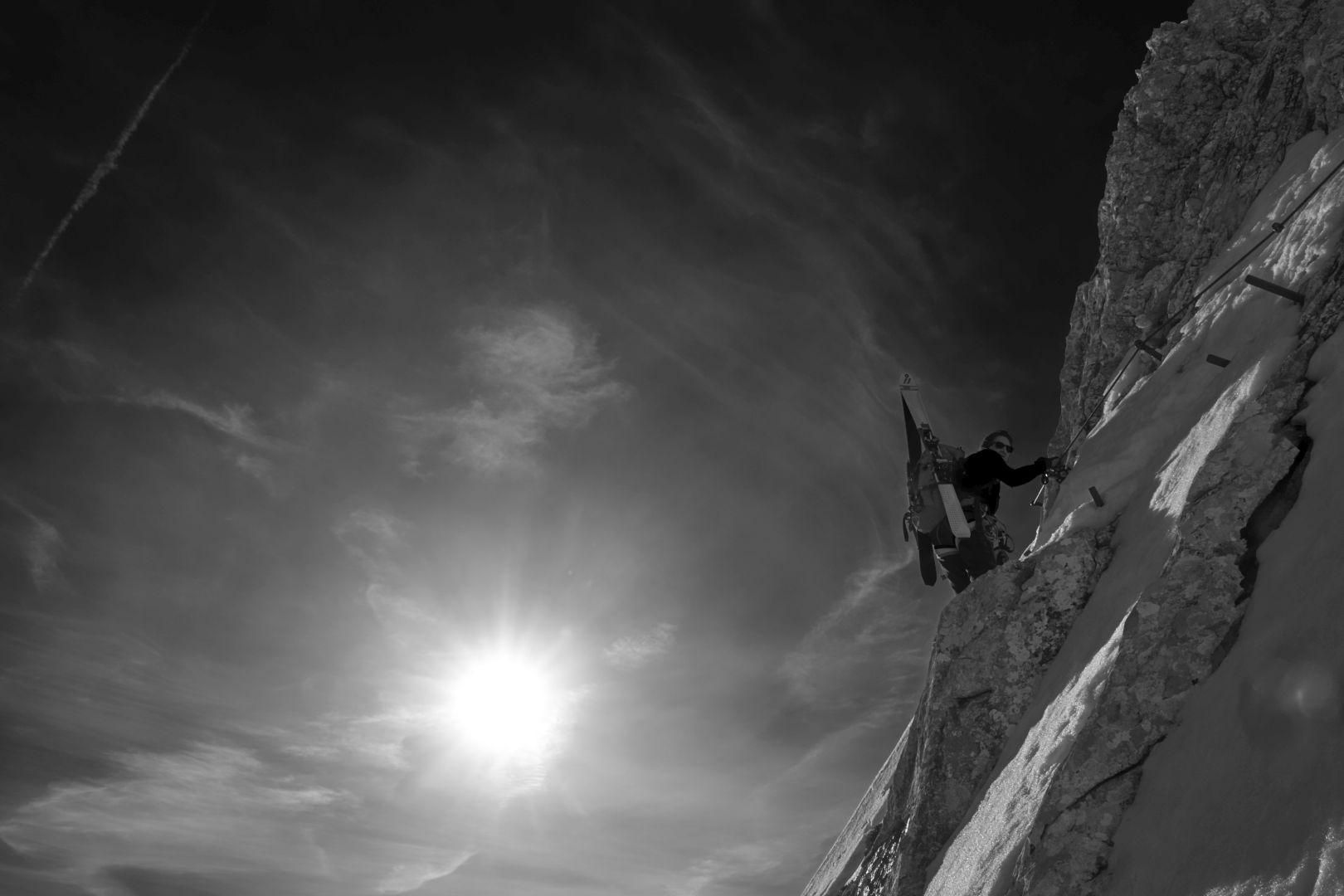 Schwarz-weiß: Bergsteiger mit Skiern am Rücken klettert steile, felsige Wand hinauf.