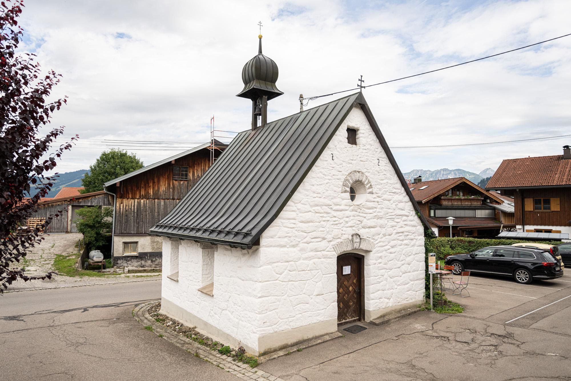 Kleine weiße Kapelle mit dunklem Dach und einem Glockentürmchen. Eine Holztür und runde Fenster sind sichtbar. Die Kapelle steht neben einer Straße und Parkplätzen.