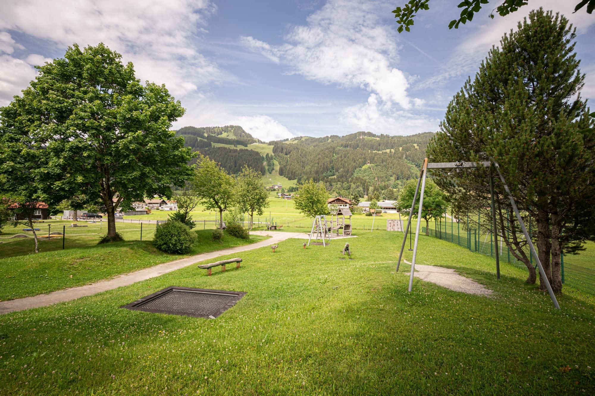 Ein grüner Park mit Bäumen und einer Schaukel steht im Vordergrund. Im Hintergrund sind Hügel und ein blauer Himmel zu sehen.