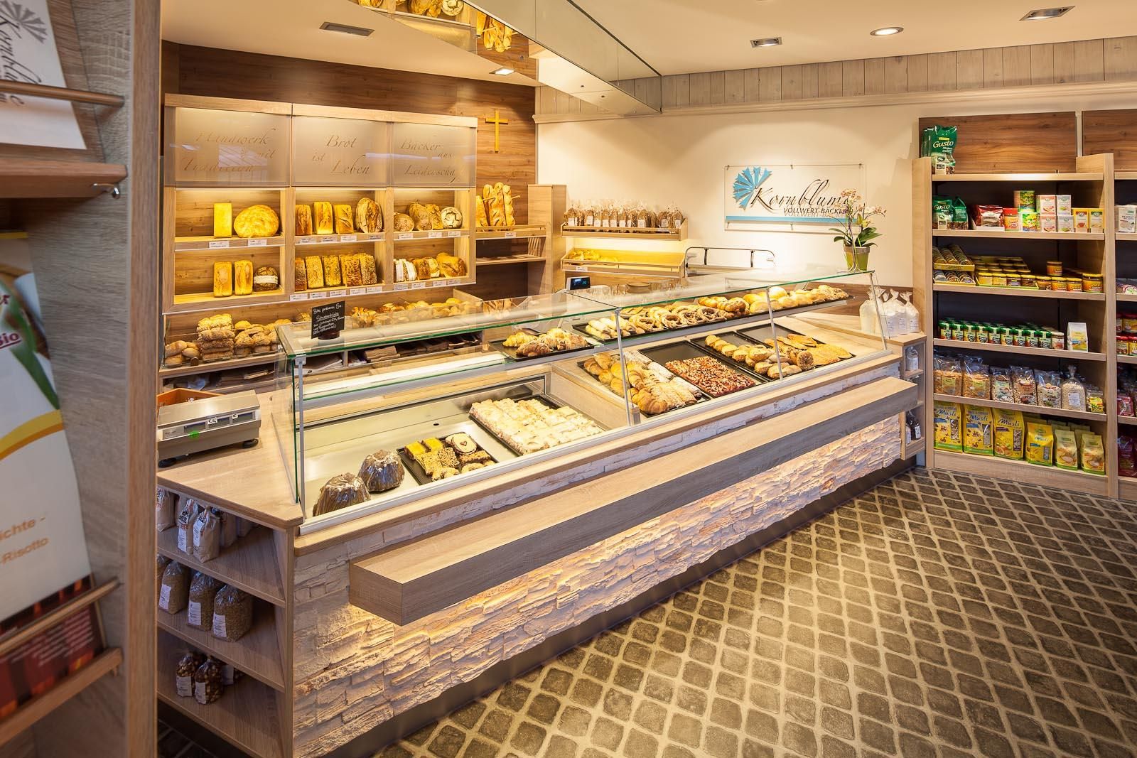 Helle Bäckerei mit Auslagen voller Backwaren. Regale mit verpackten Lebensmitteln im Hintergrund.