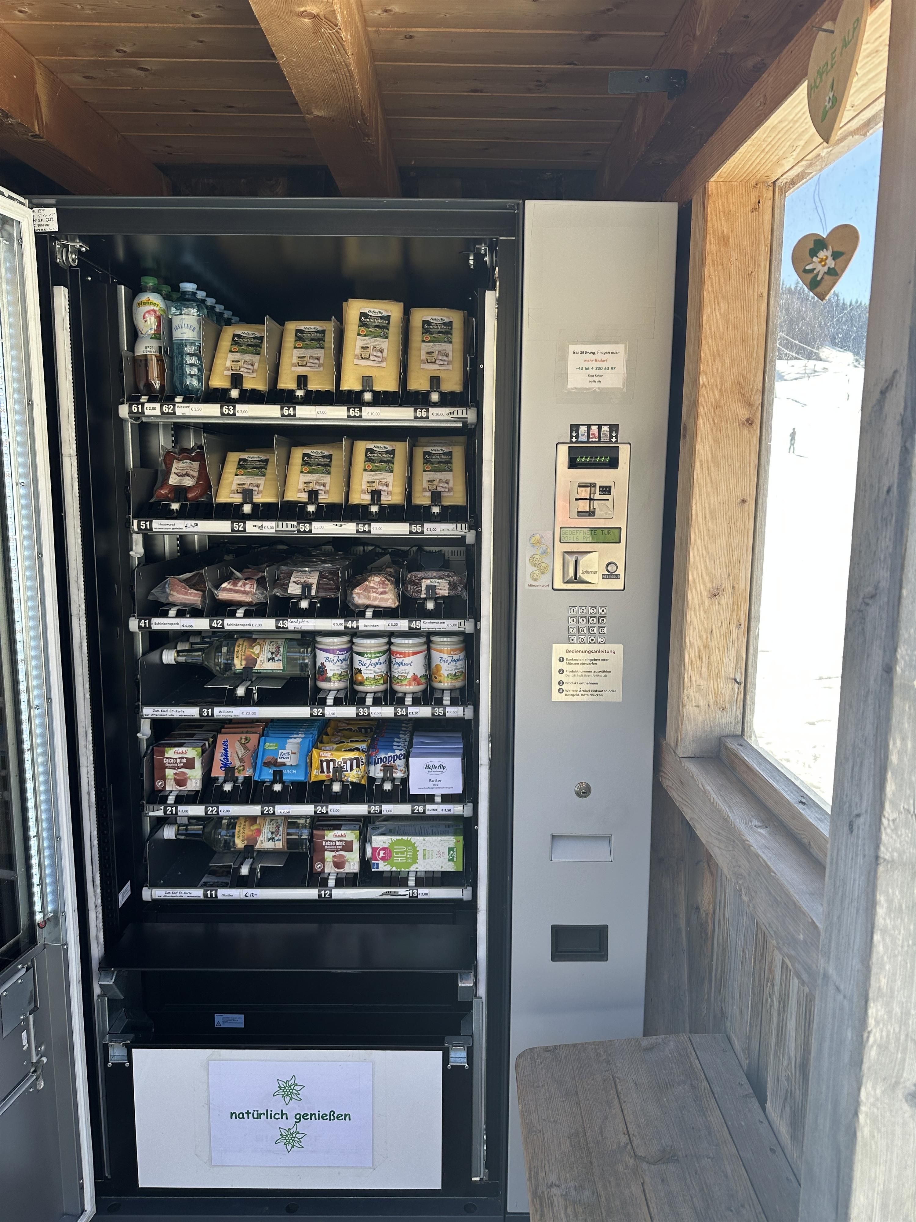 <p>Nahaufnahme des Höfle Alp Automaten. Mehrere Regale gefüllt mit Käse, Wurst, Getränken und Snacks. Beschriftungen an den Fächern.</p>