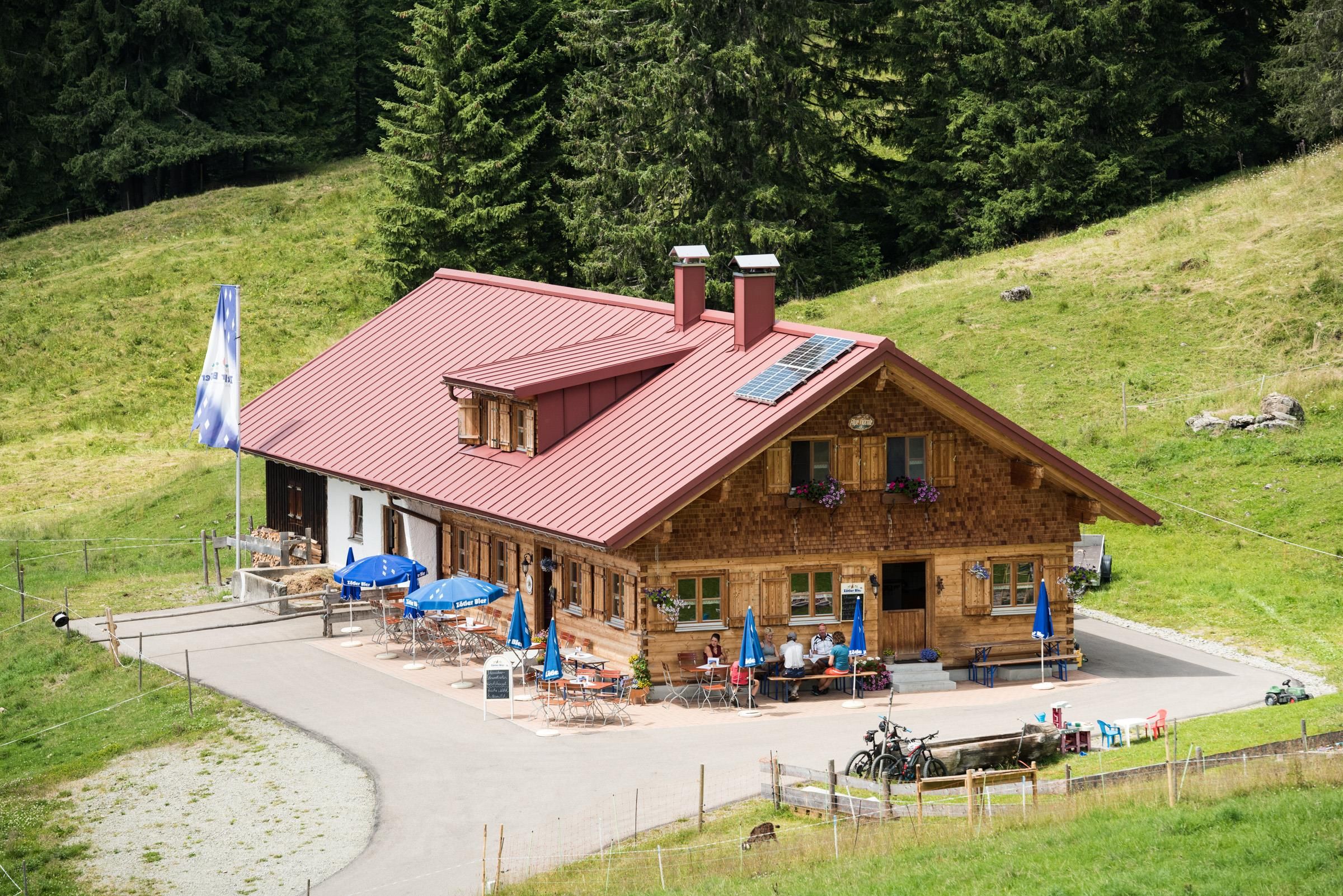 Ein hölzernes Gebäude mit rotem Dach und blauen Sonnenschirmen auf der Terrasse. Umgeben von grünen Wiesen und dunklen Bäumen an einem Hang.