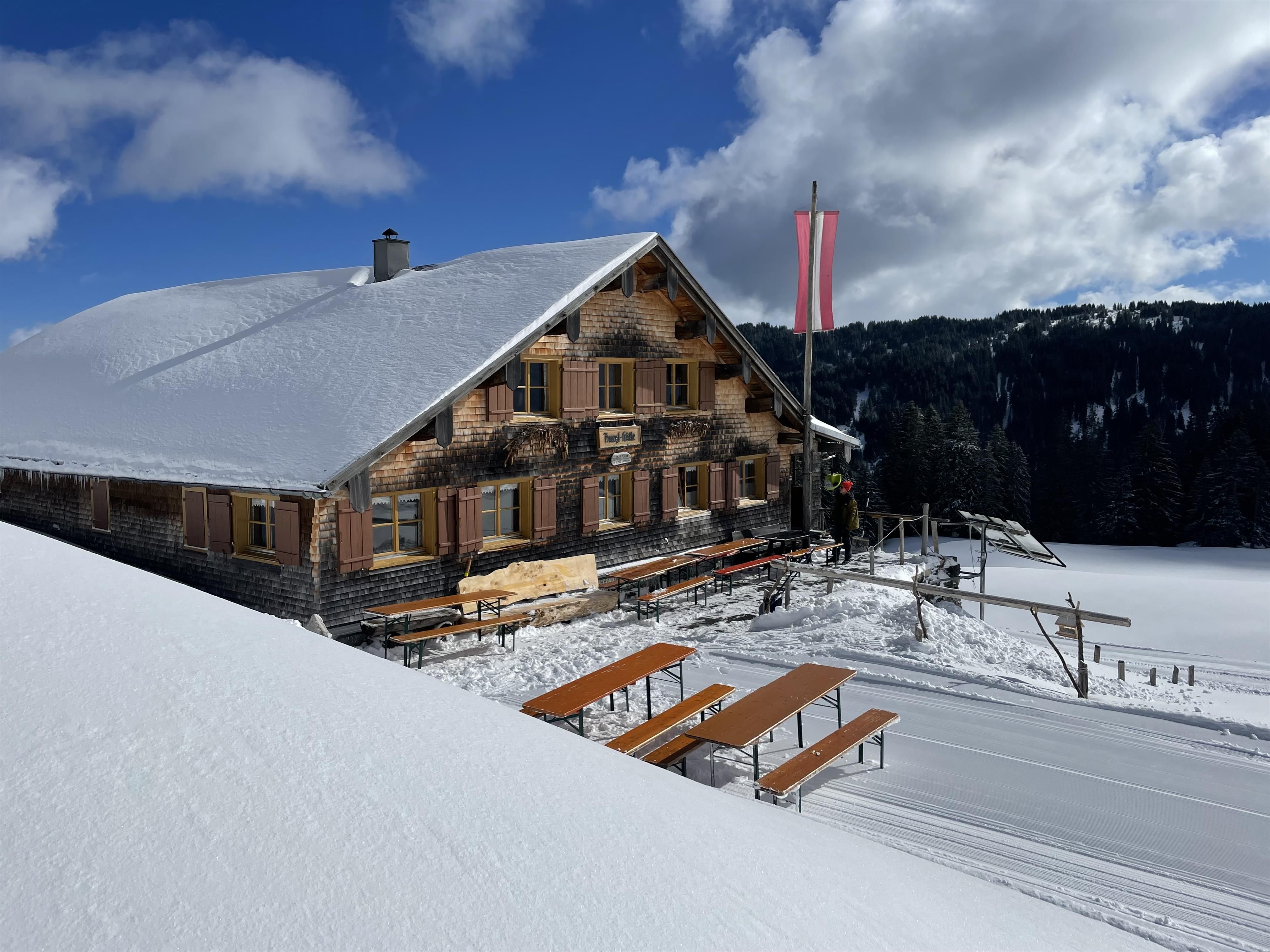 Schnee bedeckte Hütte mit Holzwänden und braunen Fensterläden. Mehrere Holztische und Bänke im Schnee.