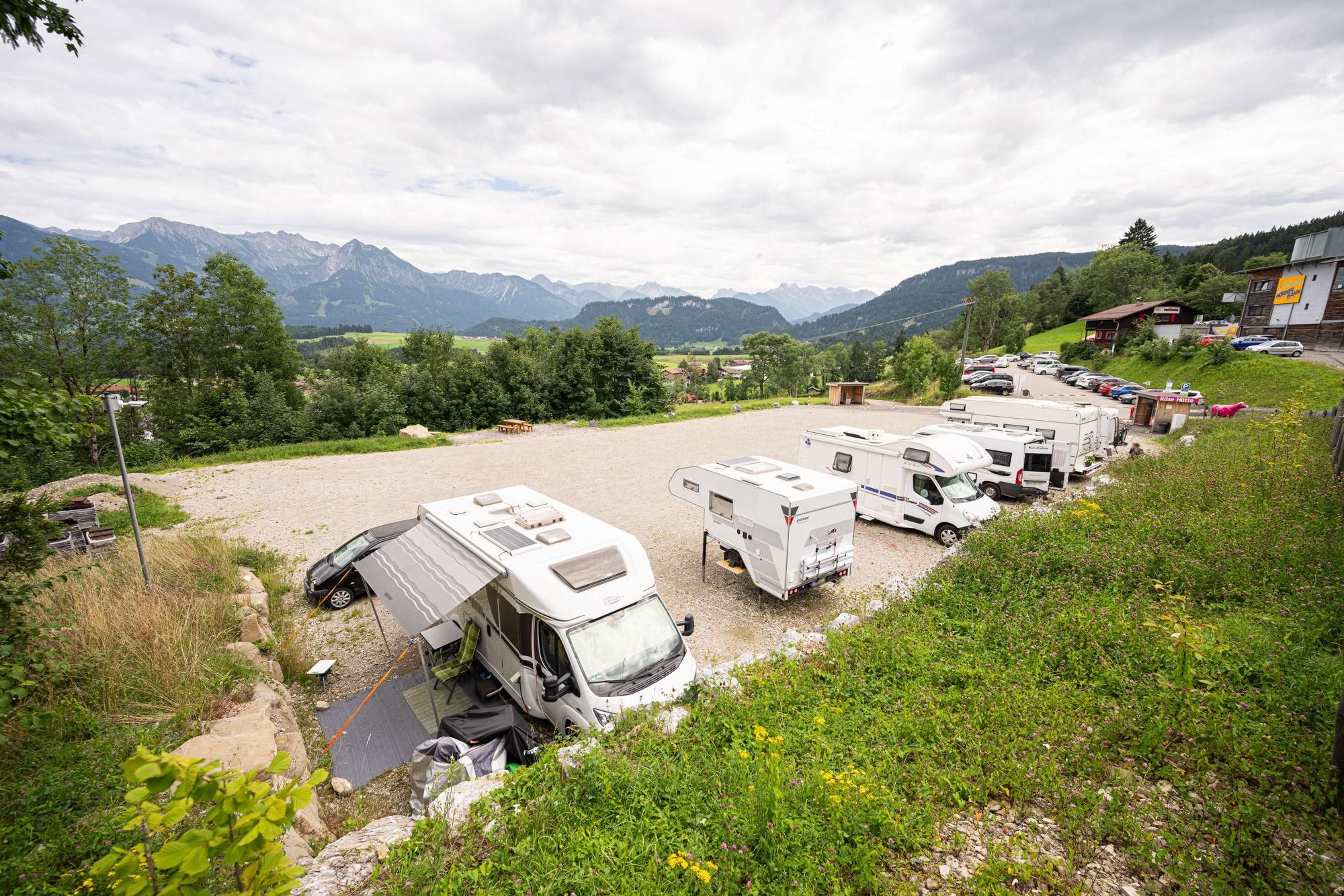 Wohnmobilstellplatz Hörnerbahn mit mehreren weißen Reisemobilen auf Schotter. Im Hintergrund grüne Landschaft und Bergpanorama unter bewölktem Himmel.