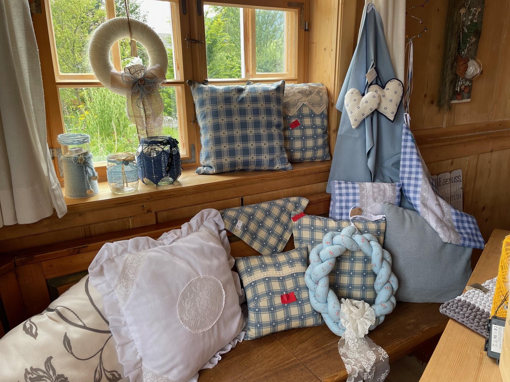 Marktstand: Dekokissen (kariert, weiß), Kränze, Gläser, Stoffherzen liegen auf einer Holzbank vor einem Fenster.