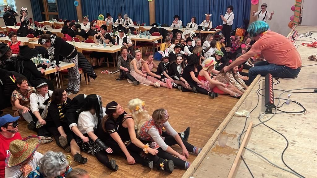 Eine lebhafte Szene einer Faschingsveranstaltung in einem großen Saal. Viele kostümierte Menschen sitzen auf dem Boden vor einer Bühne.