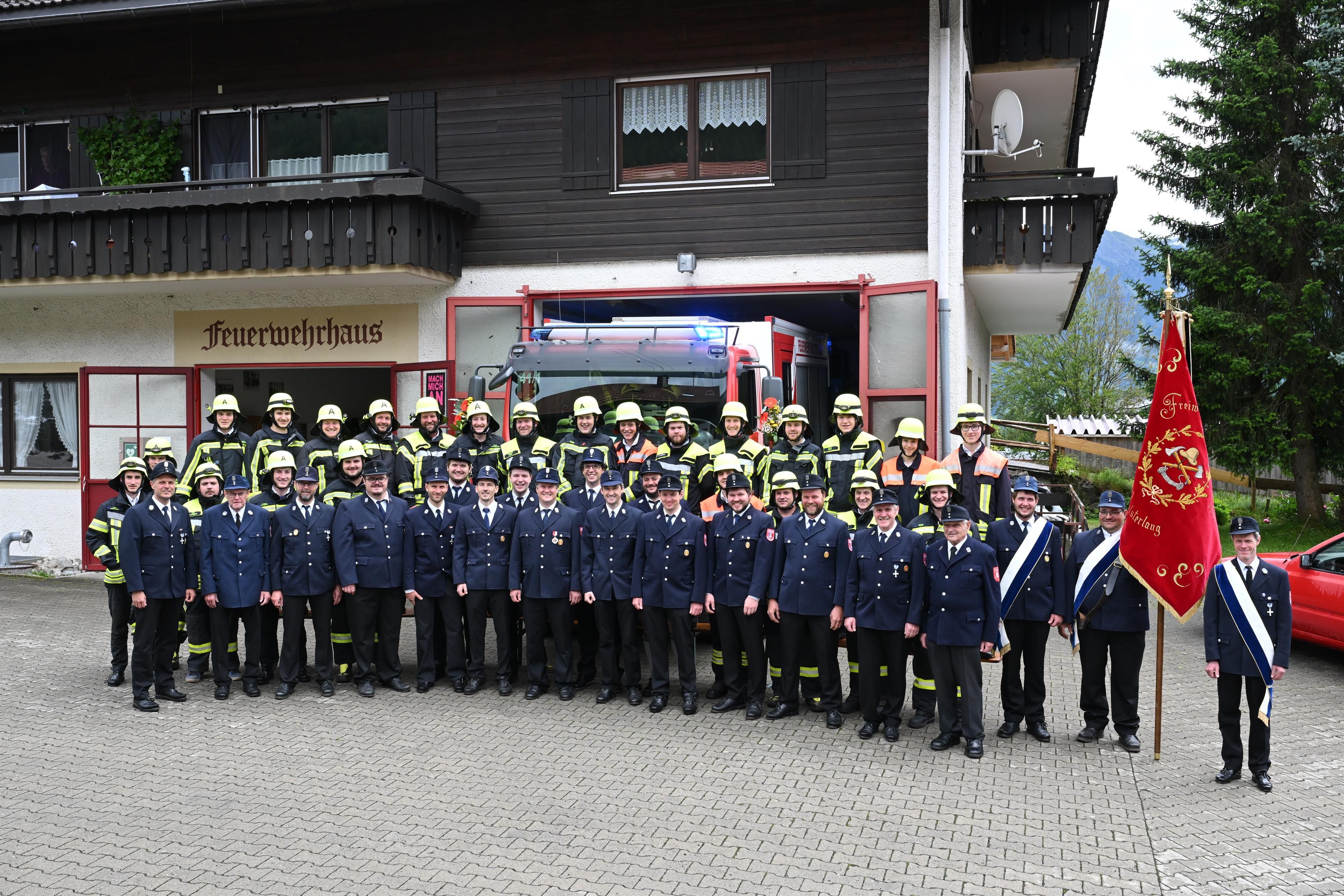 Eine Gruppe von Männern steht vor einem Haus mit der Aufschrift "Feuerwehrhaus". Die erste Reihe trägt eine blaue Uniform,  die Reihen zwei und drei Löschanzüge. Daneben wird die rote Vereinsfahne gehalten.