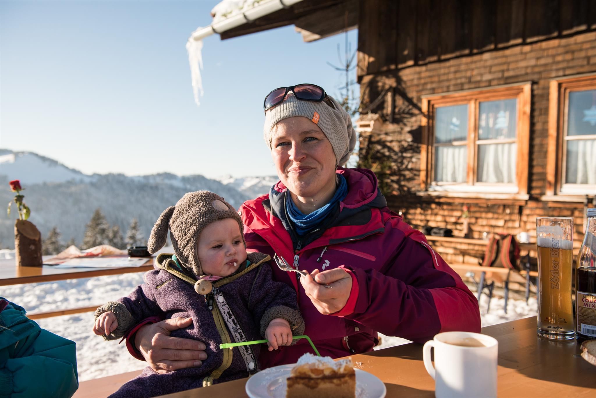 Mutter mit Kleinkind im Schnee vor Berghütte: Frau in roter Jacke hält Kind mit Mütze, Kuchen, Kaffee, Winterlandschaft
