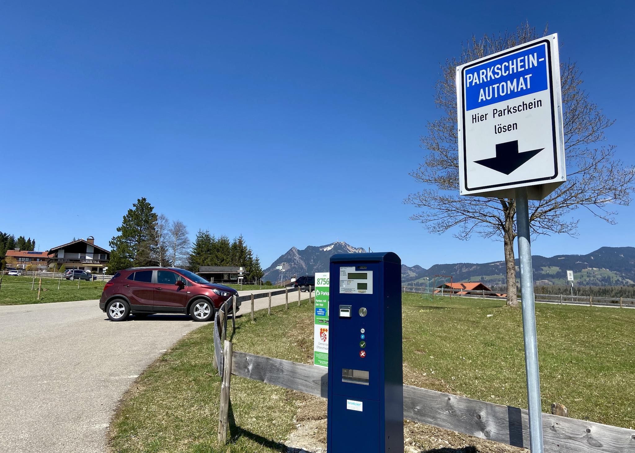 Blau-weißer Parkscheinautomat neben einem Schild "PARKSCHEIN-AUTOMAT Hier Parkschein lösen" mit Pfeil nach unten