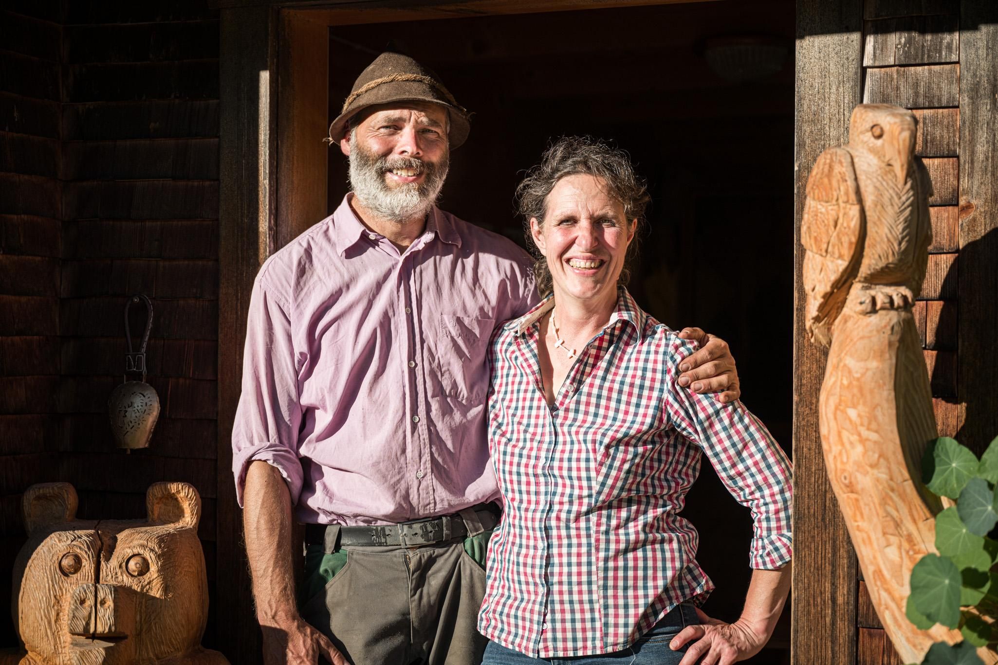 Ein Mann und eine Frau stehen freundlich vor einem Holzgebäude. Im Hintergrund sind kunstvoll geschnitzte Holzfiguren zu sehen.