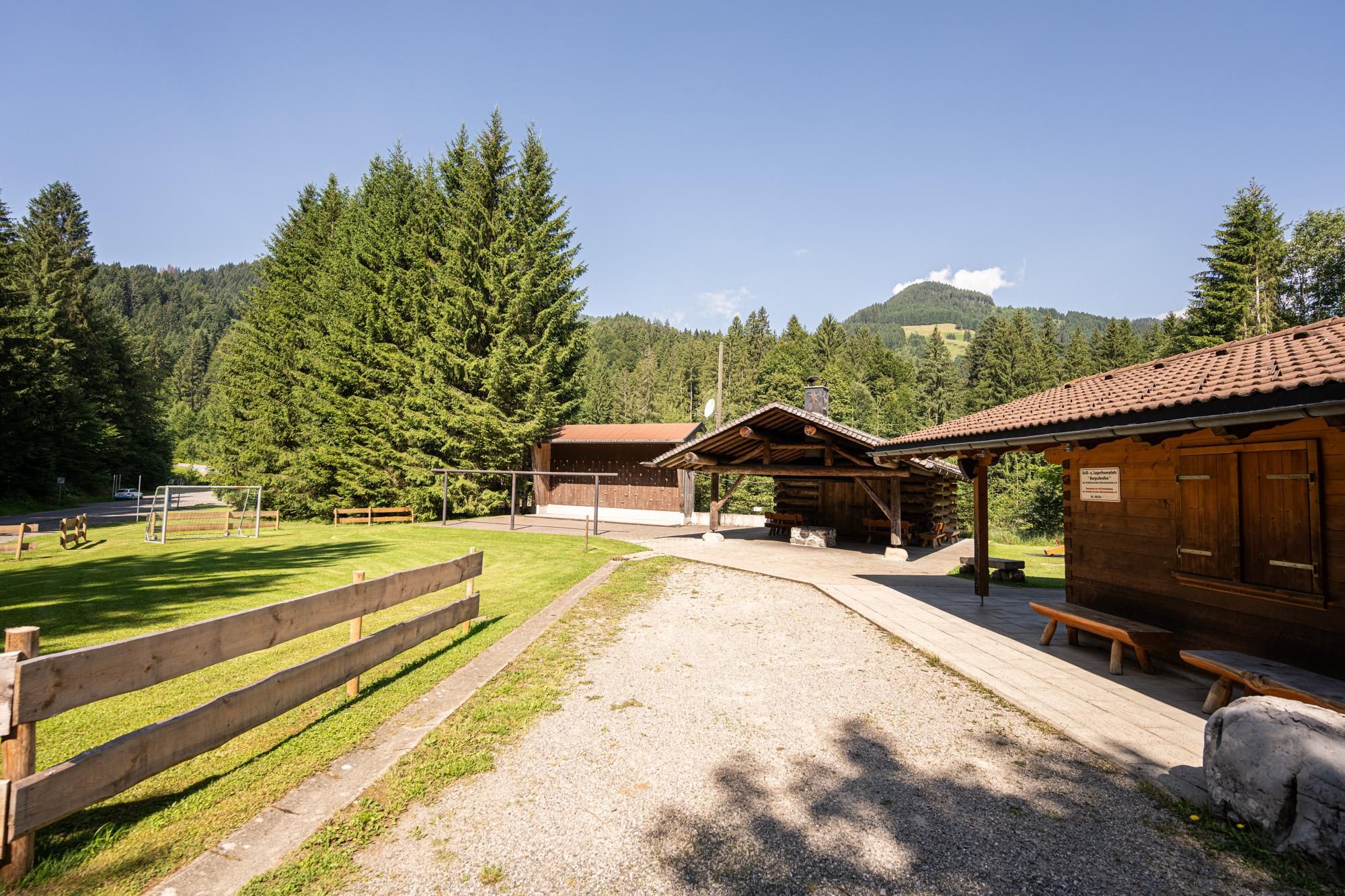 Ländlicher Grillplatz "Burgschrofen" in Obermaiselstein. Eine Holzscheune und eine offene Holzhütte mit Kamin stehen auf einer grünen Wiese.