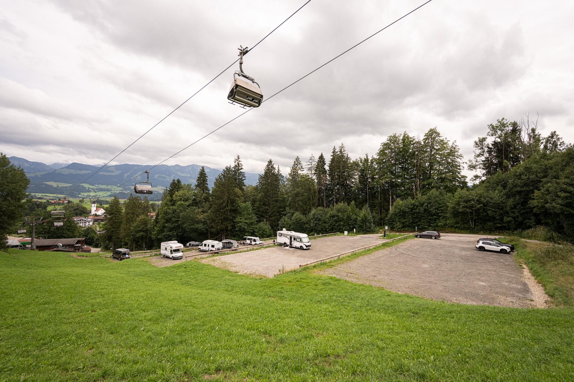 Parkplatz am Weltcup-Express Ofterschwang mit mehreren Wohnmobilen und PKWs. Darüber eine Seilbahn mit zwei Sesseln vor bewaldetem Hang und bewölktem Himmel.