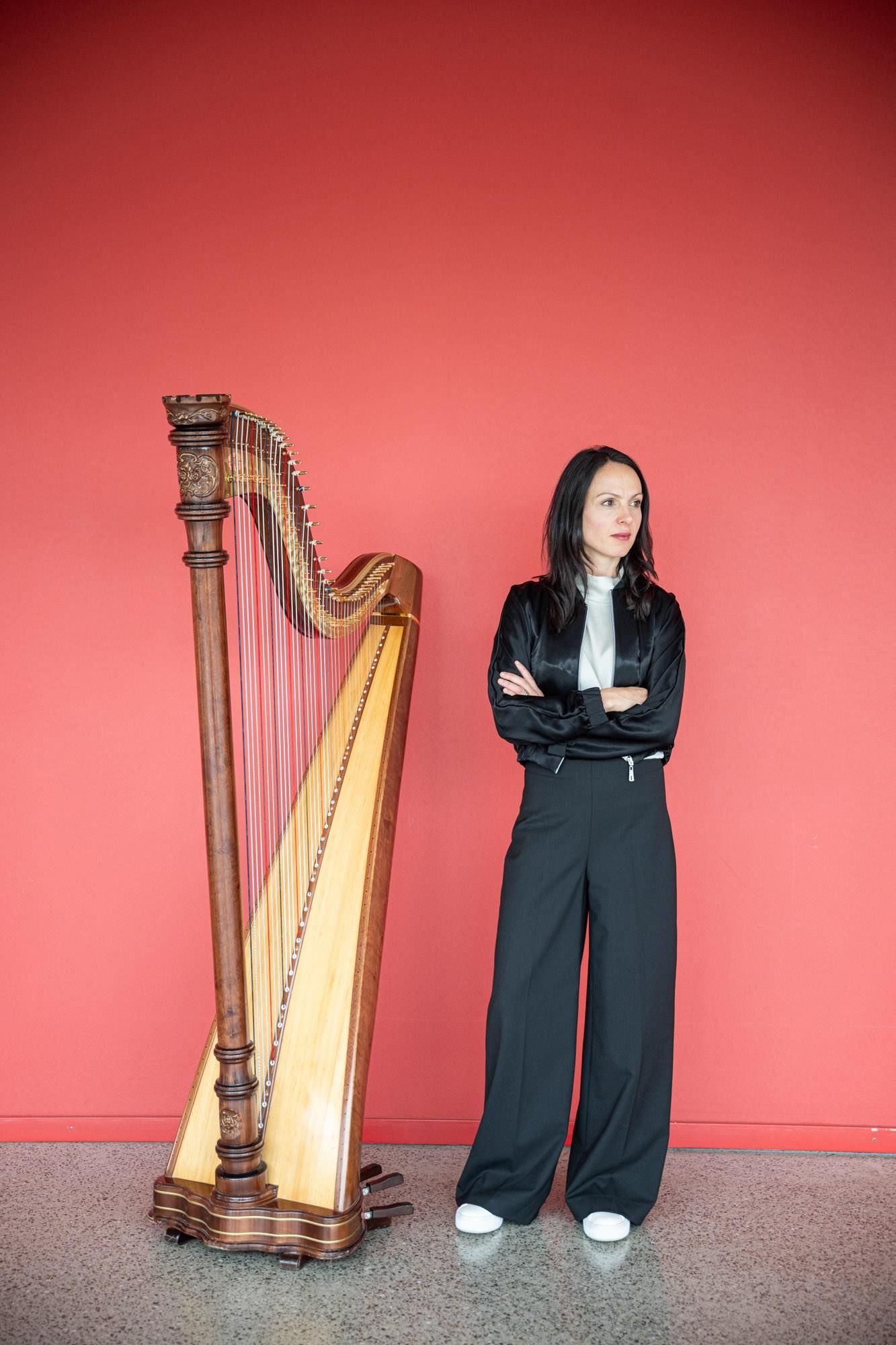Die Musikerin Melinda Rodriguez positioniert sich neben ihrer Harfe stehend in schwarzem Anzug und weißer Bluse im Fotostudio vor roter Leinwand.