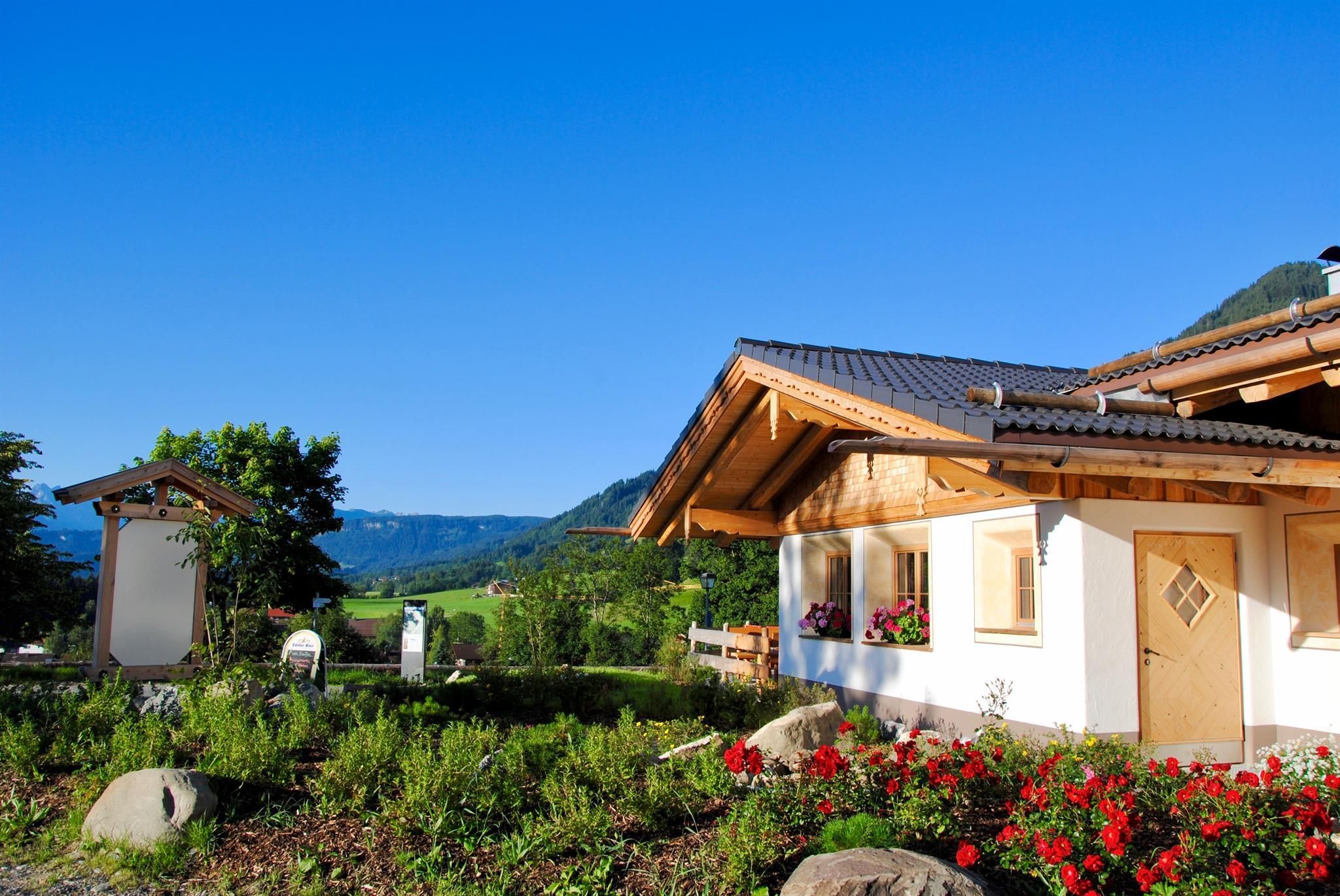 Helles Holzhaus mit dunklem Dach und weißen Wänden, umgeben von grüner Vegetation, roten Blumen.