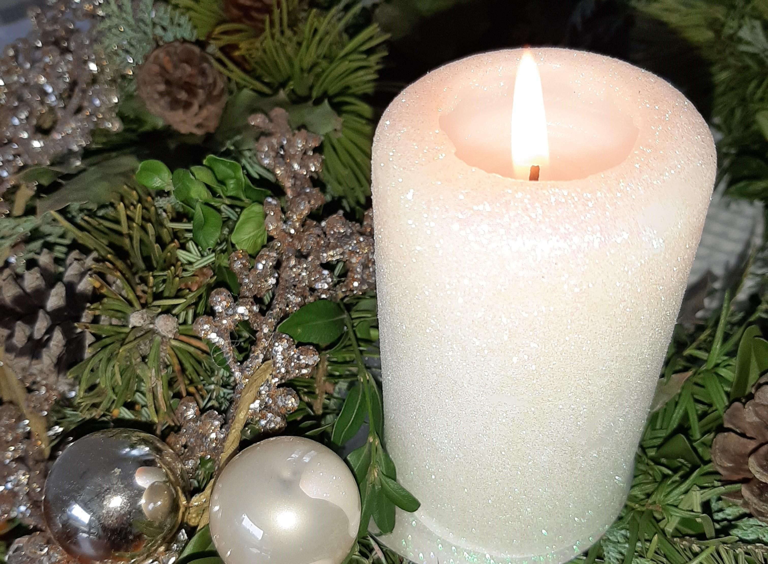 Ein Adventsgesteck mit weißer, brennender Kerze, silberner und weißer Dekoration und Tannenzweigen.