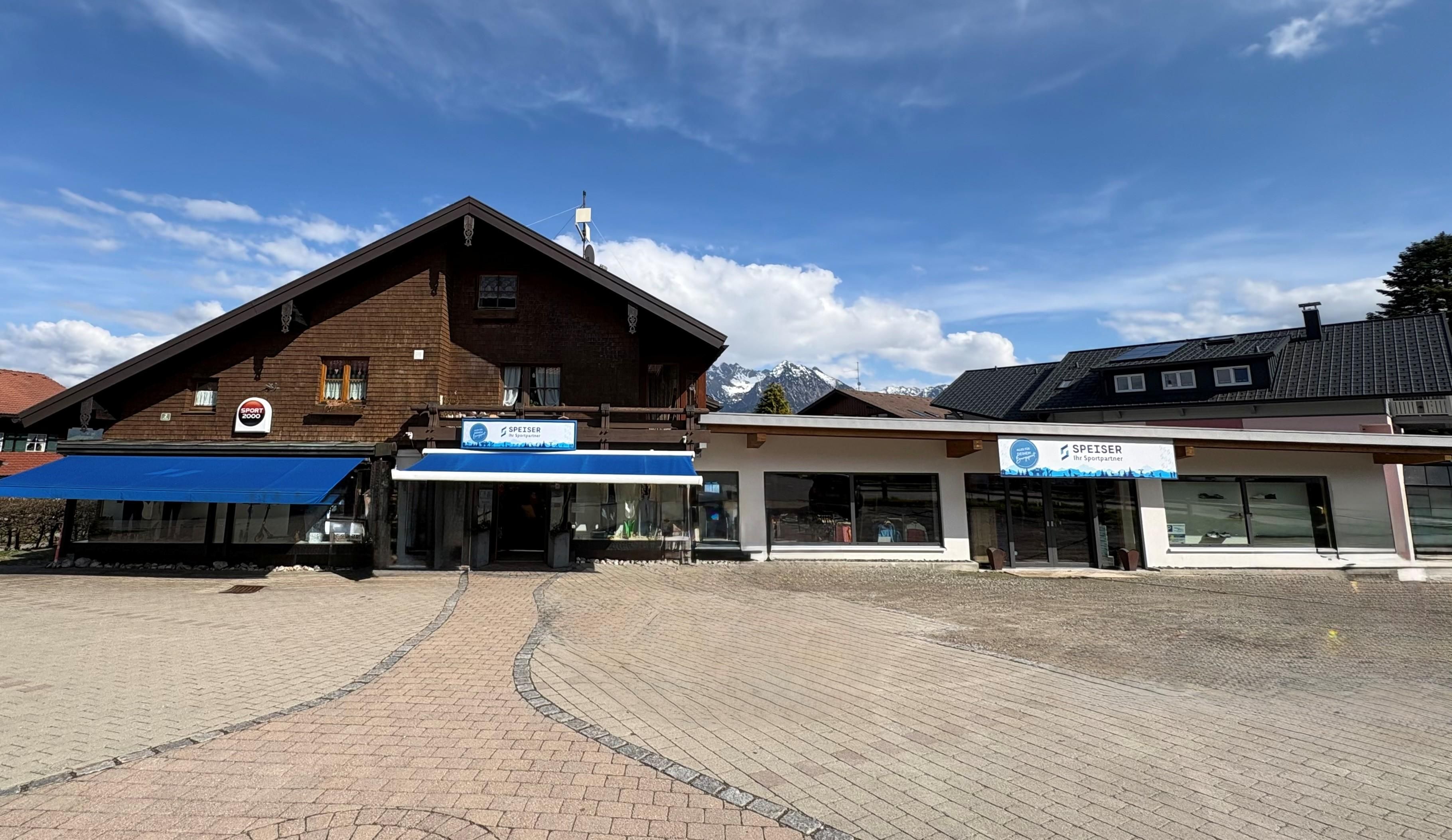 Ein modernes Geschäftsgebäude mit einem traditionellen Dach in einer malerischen Umgebung. Im Vordergrund ist ein gepflasterter Platz sichtbar.
Seit 2 Gerarationen Familienbetrieb Sport Shop Speiser Bolsterlang.