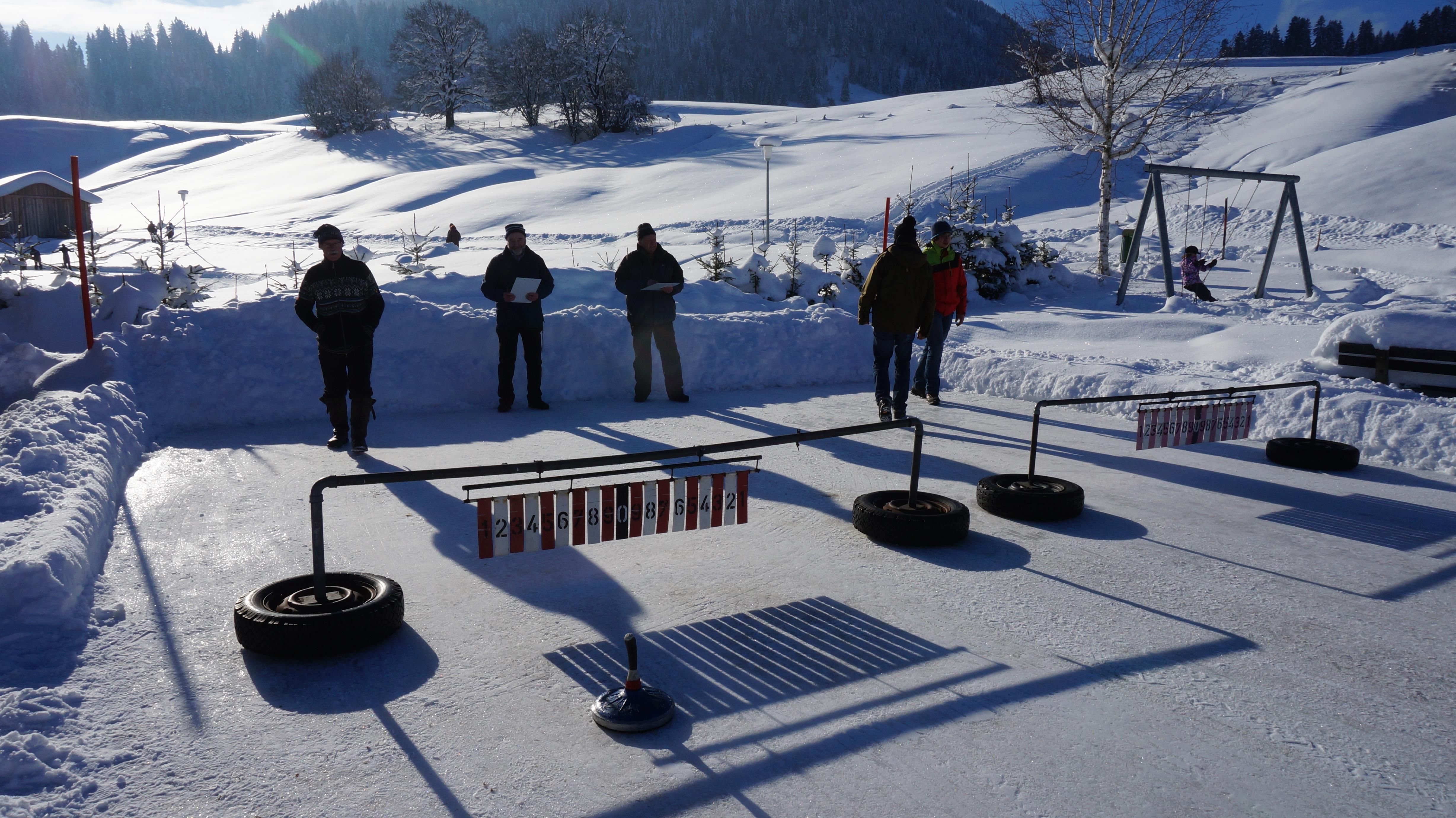 Auf einer verschneiten Eisstockbahn stehen mehrere Personen und schauen zu. Im Vordergrund sind Eisstöcke und Zielringe zu sehen.