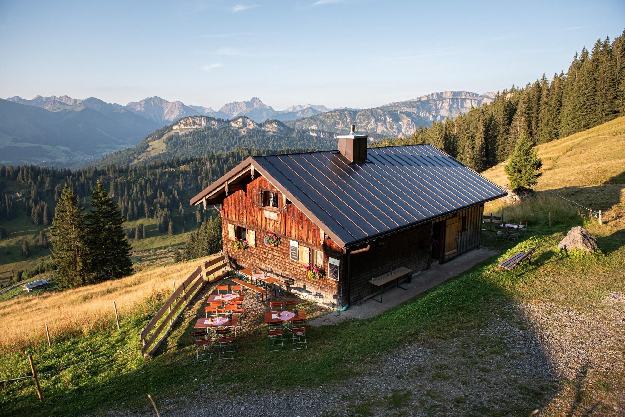 Eine charmante Alphütte in den Bergen mit Holzwänden und einem Blechdach. Im Vordergrund sind Tische auf einer Wiese und die umliegenden Berge sind sichtbar.