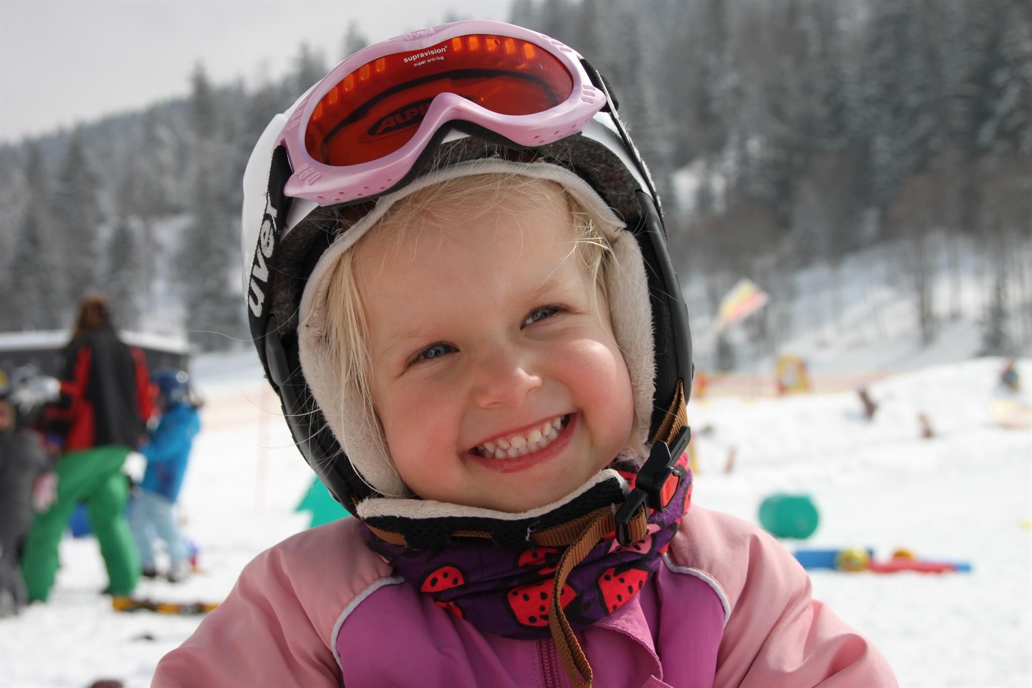 Lächelndes Kind mit blonden Haaren, Skihelm und pinkfarbener Skibrille, im Hintergrund verschneite Landschaft.