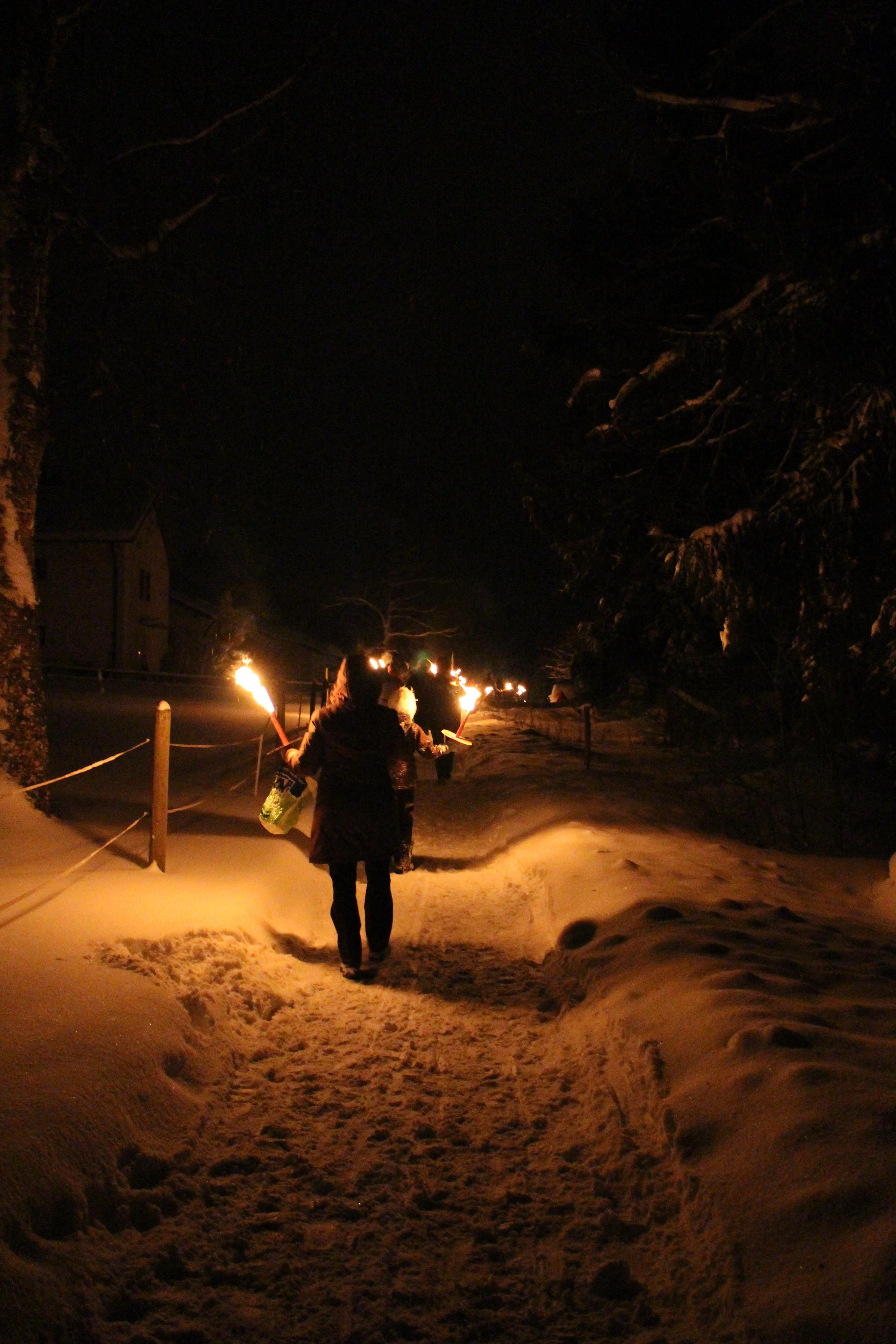 Auf einem Weg, der mit Schnee bedeckt ist, läuft eine Gruppe mit Laternen und Fackeln in der Nacht.