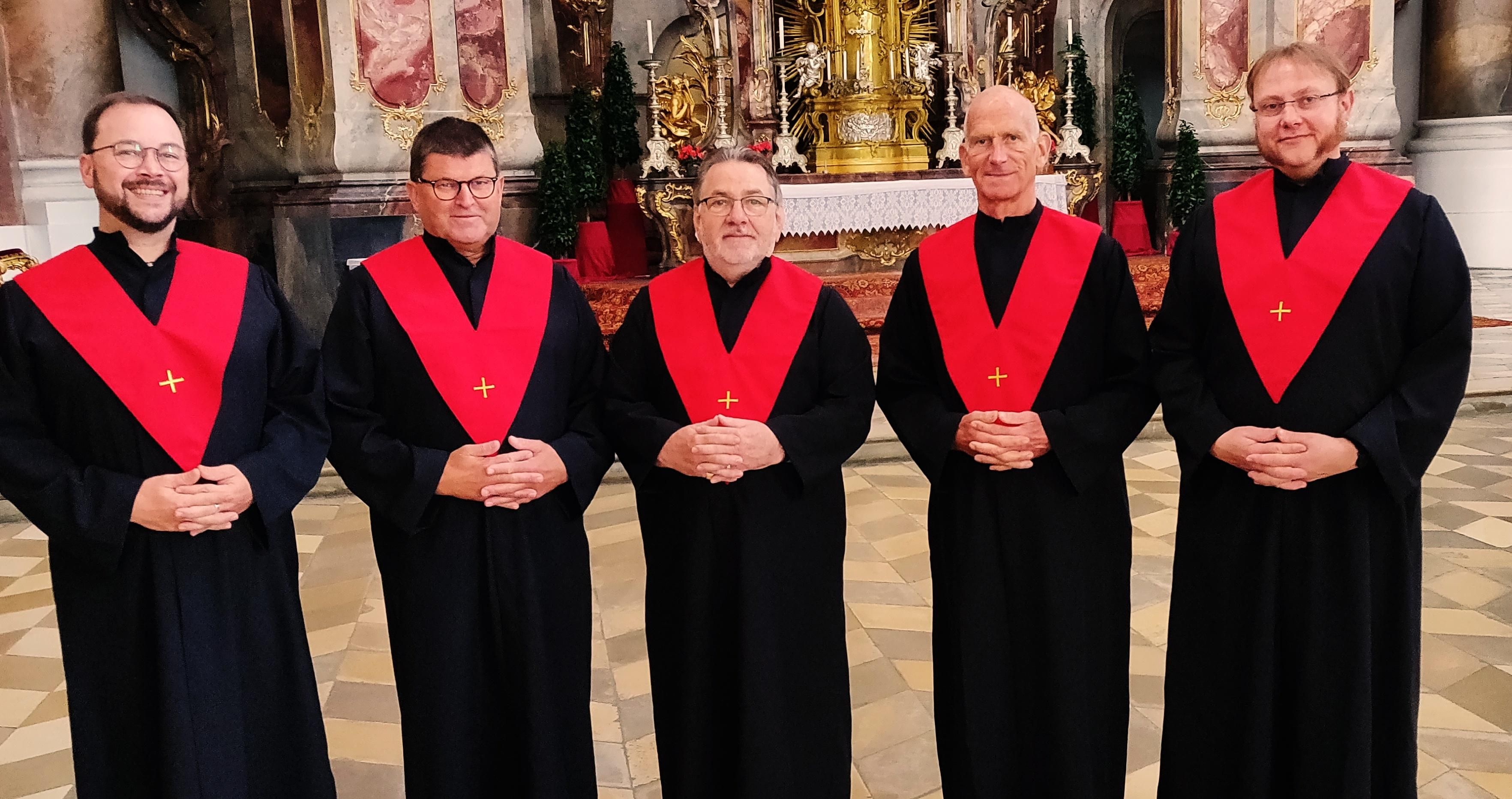Der Chor Schola Cantorum - fünf Männer, schwarze Auftrittsgewänder mit rotem Schal, darauf ein gesticktes Kreuz.