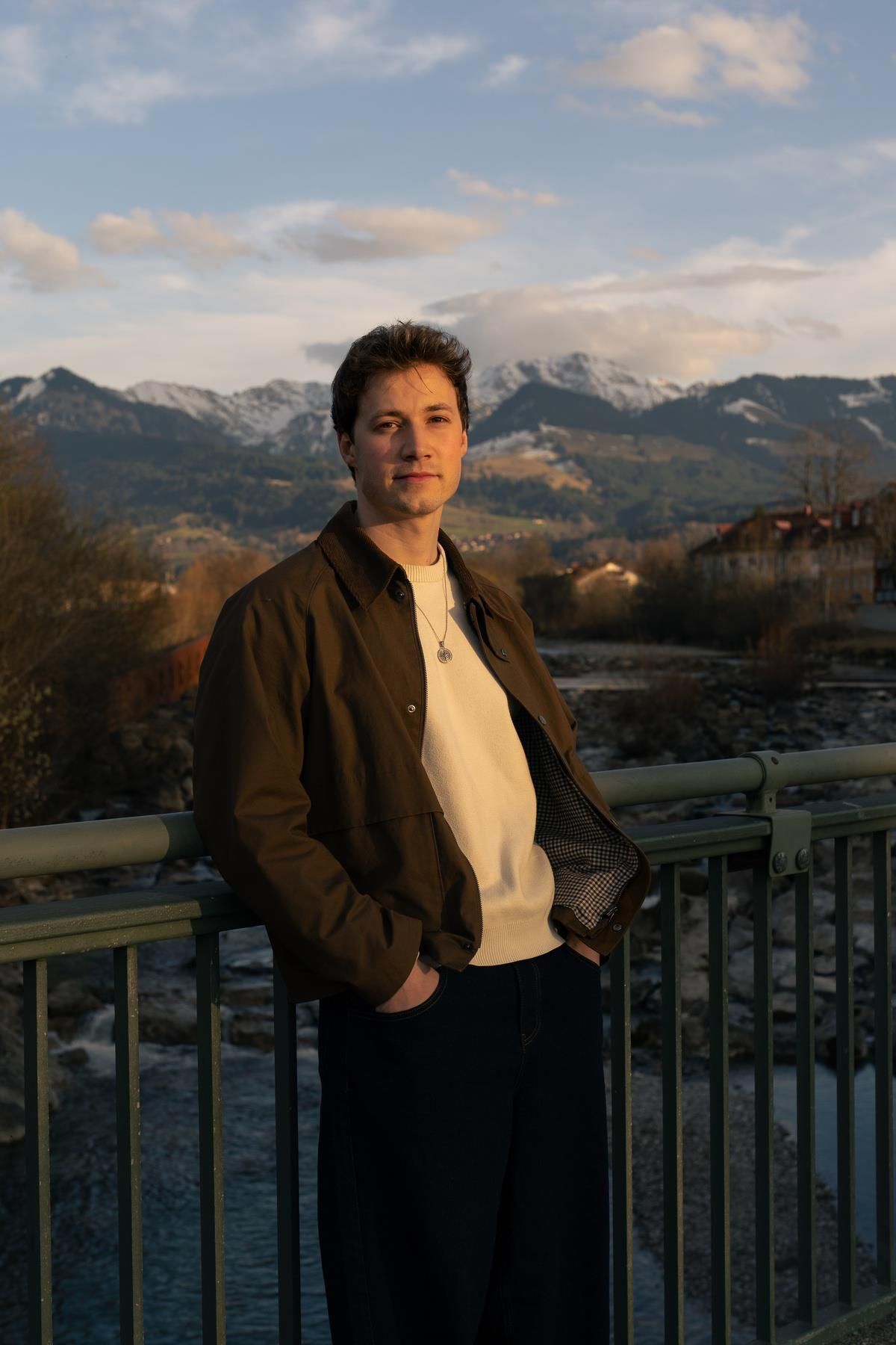 Flo Wurm ist jung, hat braune Haare, er steht in brauner Lederjacke und weißem Shirt vor einer Bergkulisse im Oberallgäu.