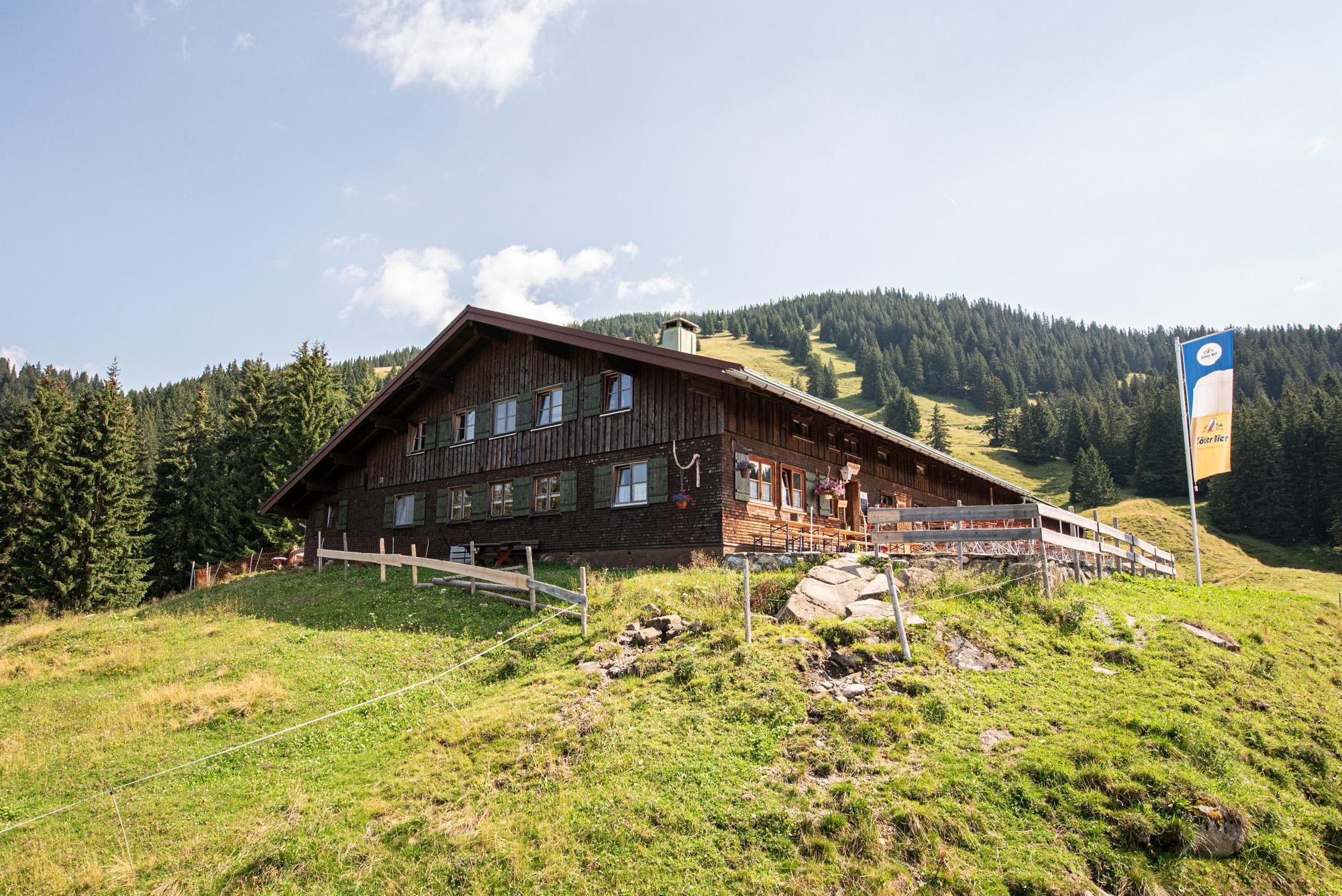 Ein rustikales Holzhaus umgeben von grünen Wiesen und Bergen. Im Hintergrund sind bewaldete Hänge und ein blauer Himmel zu sehen.