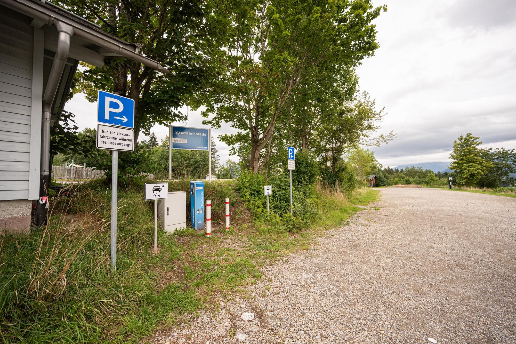 Eine E-Ladestation mit zwei blauen Säulen und einem weißen Kasten neben einem Parkplatzschild mit blauem "P". Der Untergrund ist Schotter, umgeben von Grün.