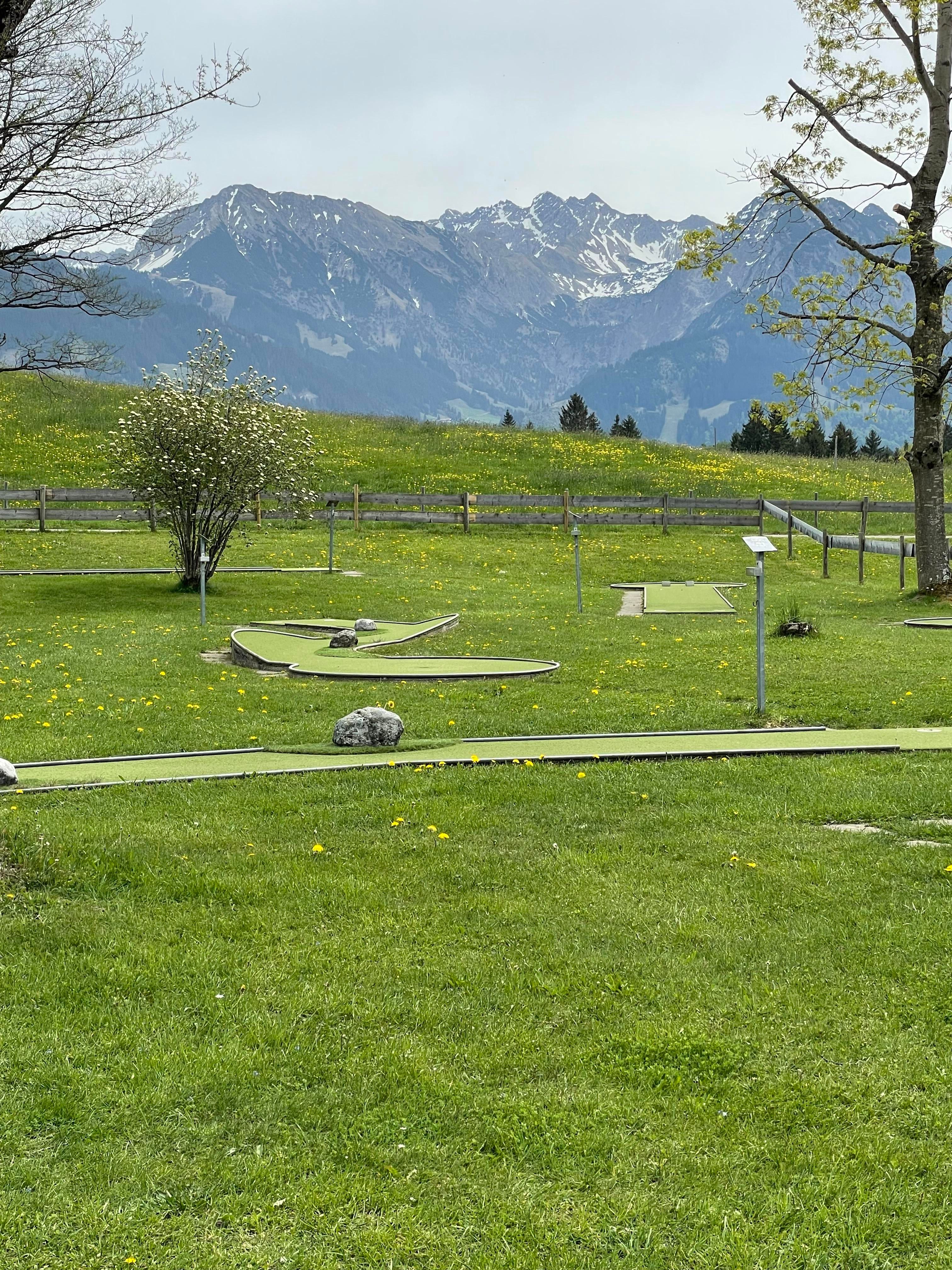 Eine Minigolfanlage in einer grünen Wiese mit Blick auf schneebedeckte Berge. Der Himmel ist bewölkt und die Landschaft ist ruhig und einladend.