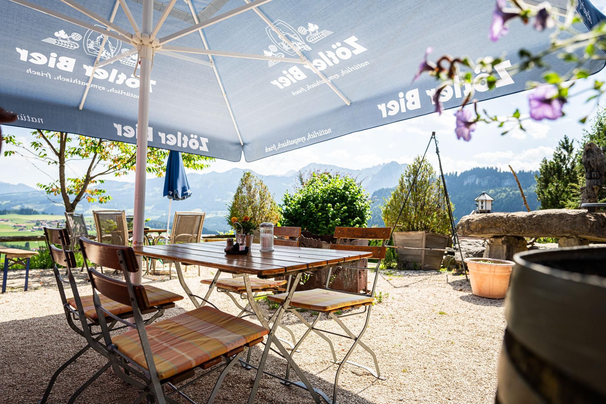 Außenterrasse des Restaurants Berghof Schwarzenberg mit Tischen und Stühlen unter großen Sonnenschirmen. Blick auf grüne Hügel und Berge unter einem hellen Himmel.
