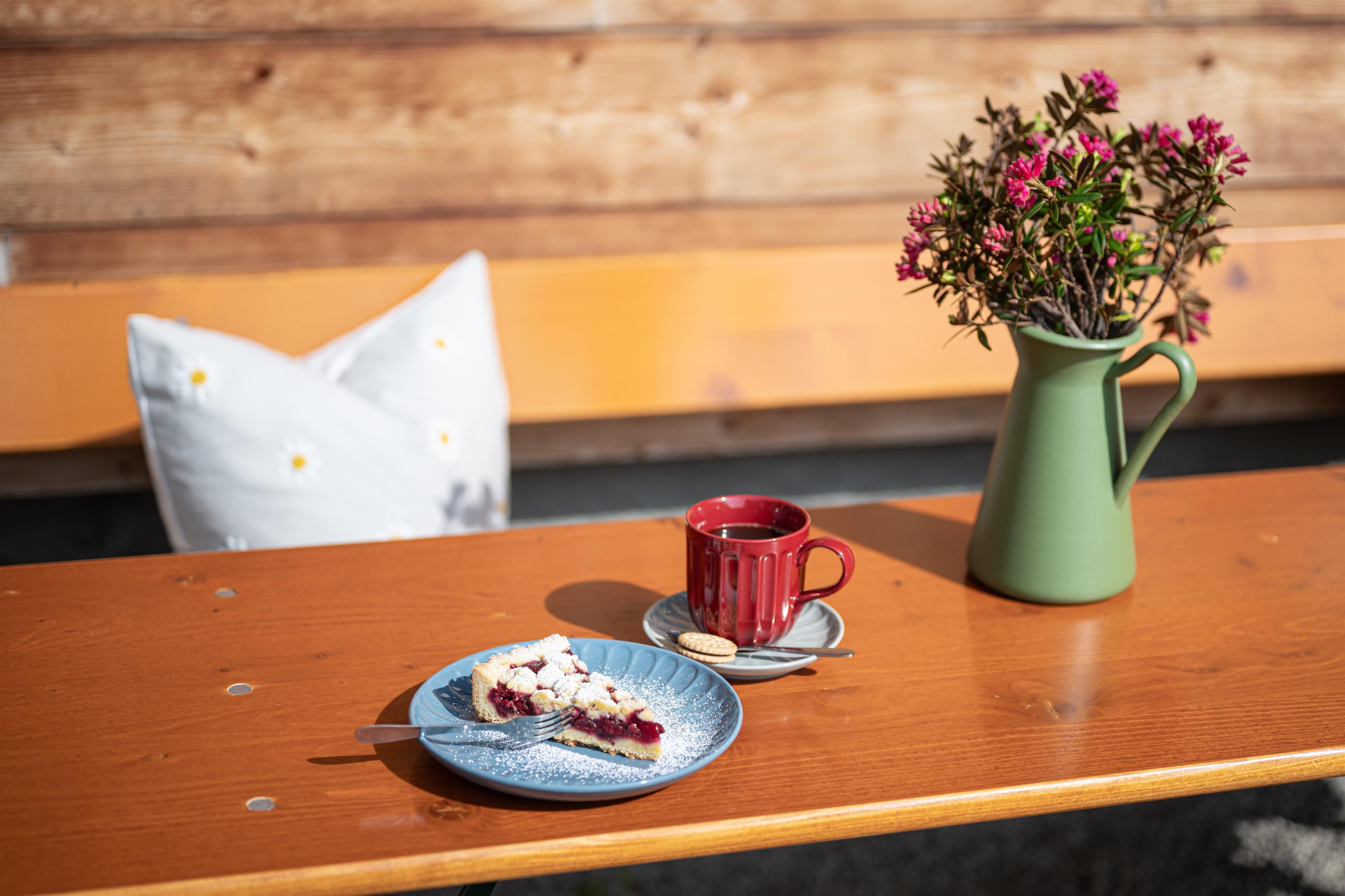 Auf einem Biertisch steht ein Stück Fruchkuchen mit Gabel. Daneben ist eine schöne rote Tasse mit Kaffee auf einem blauen Untersetzer. Auf dem Tisch steht auch eine grüne Kanne mit rosanen Wildblumen.