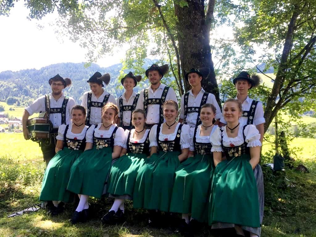 Gruppe in Tracht: 6 Männer und 5 Frauen sitzen im Grünen unter einem Baum.