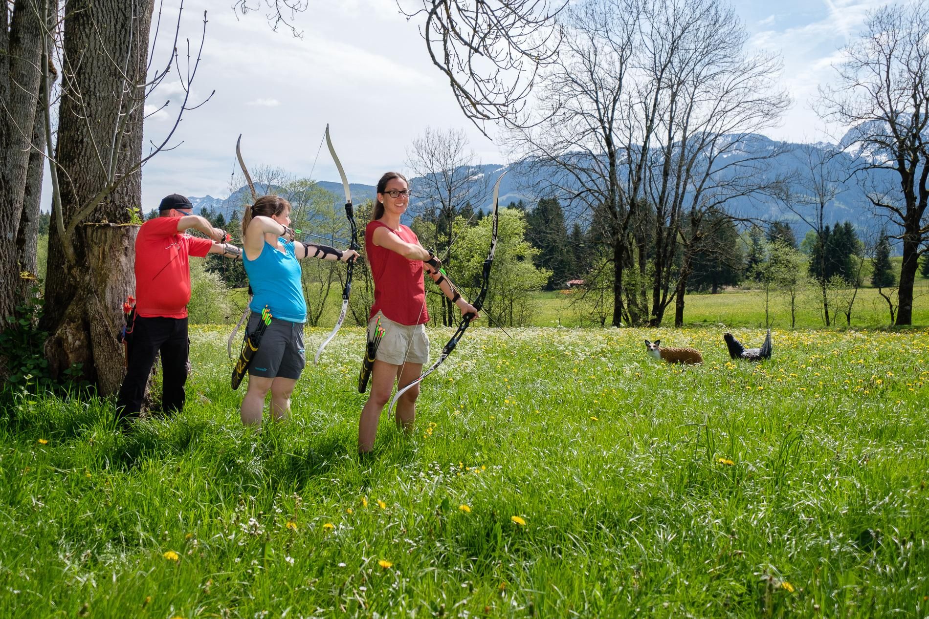 Vier Bogenschützen, zwei Männer und zwei Frauen, zielen in einer grünen Wiese mit Bergen im Hintergrund.