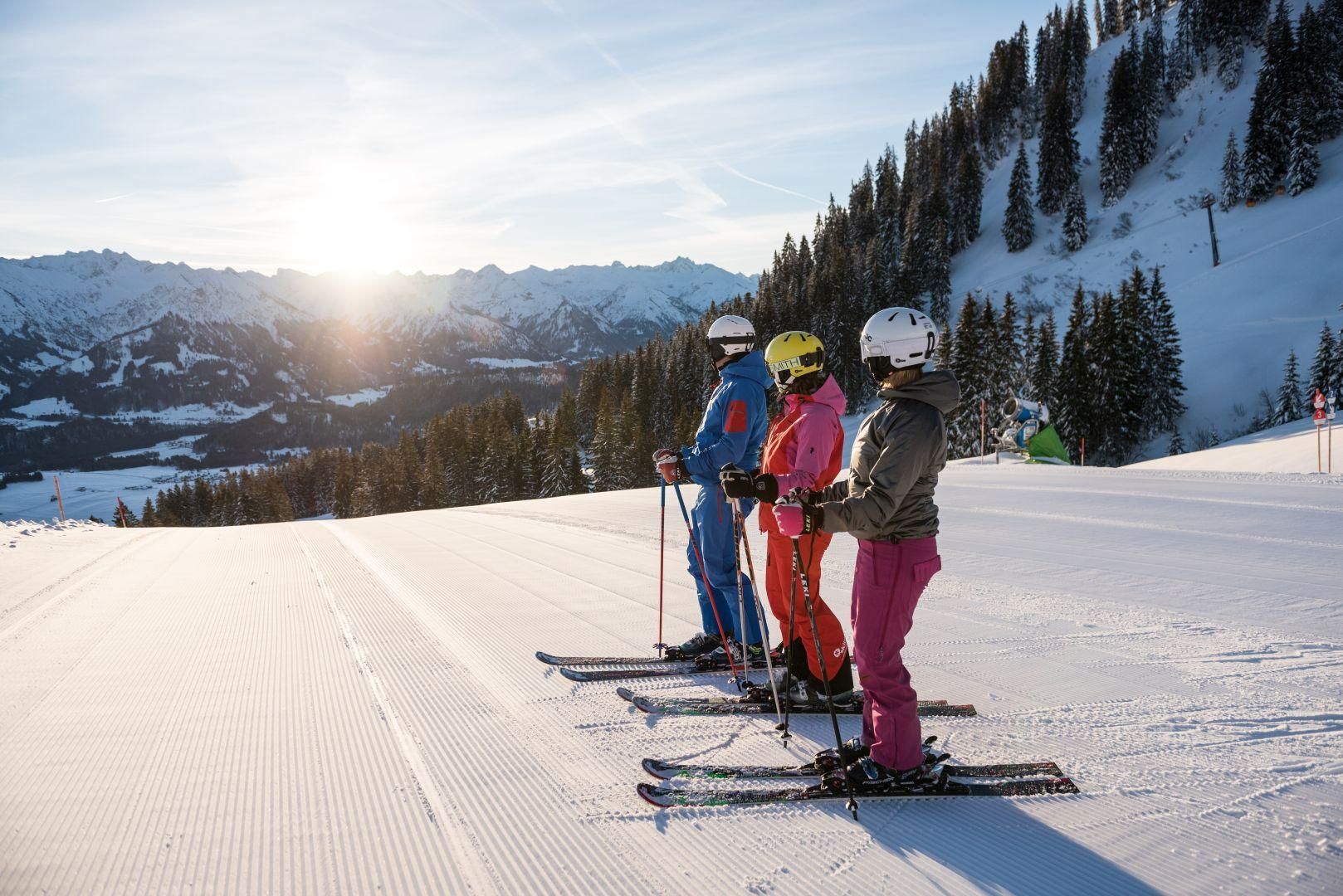 Drei Skifahrer stehen nebeneinander auf einer präparierten Skipiste. Verschneite Berglandschaft und bewaldeter Hang im Hintergrund.