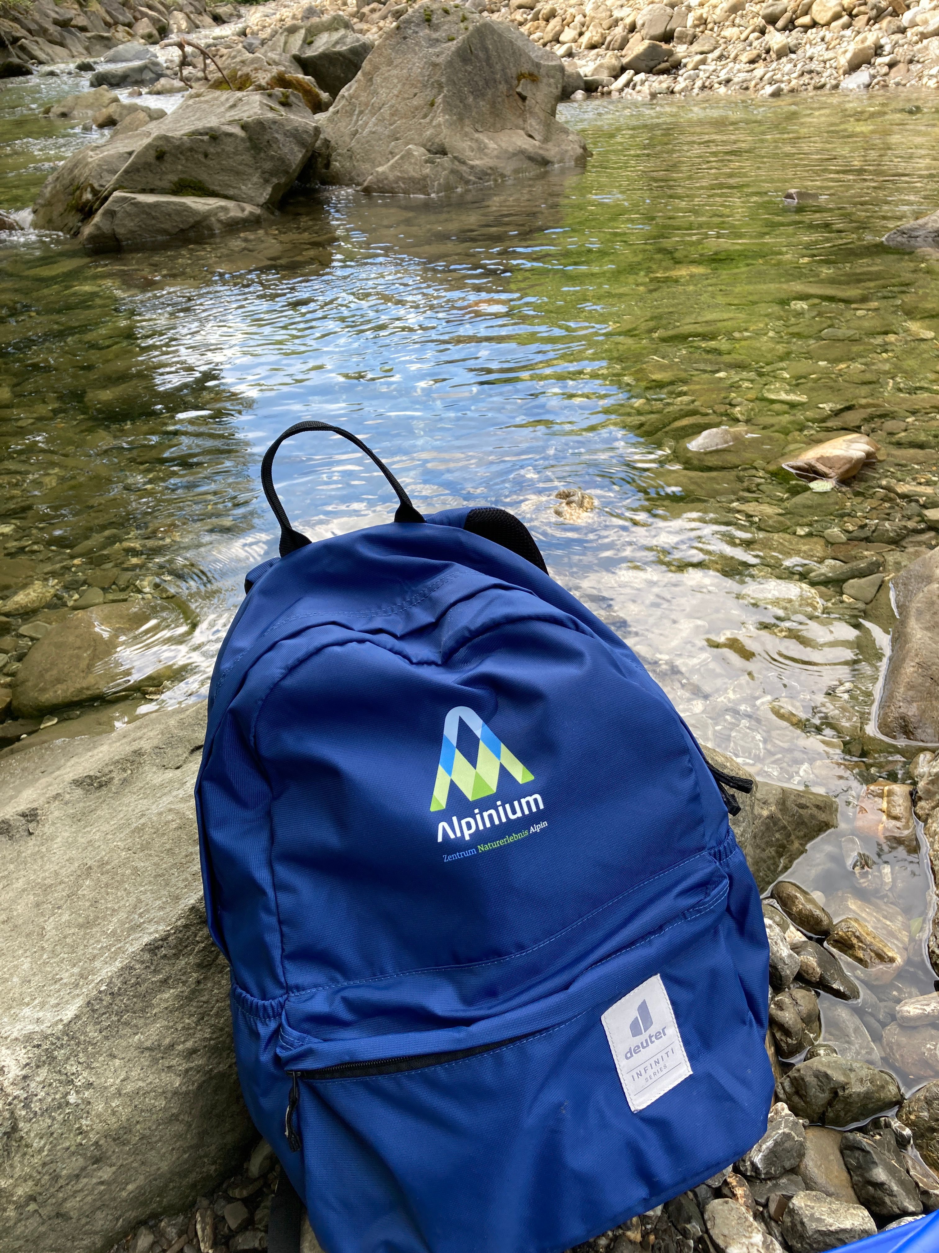 Ein blauer Rucksack mit der Aufschrift Alpinium steht auf Steinen unmittelbar am Bach.