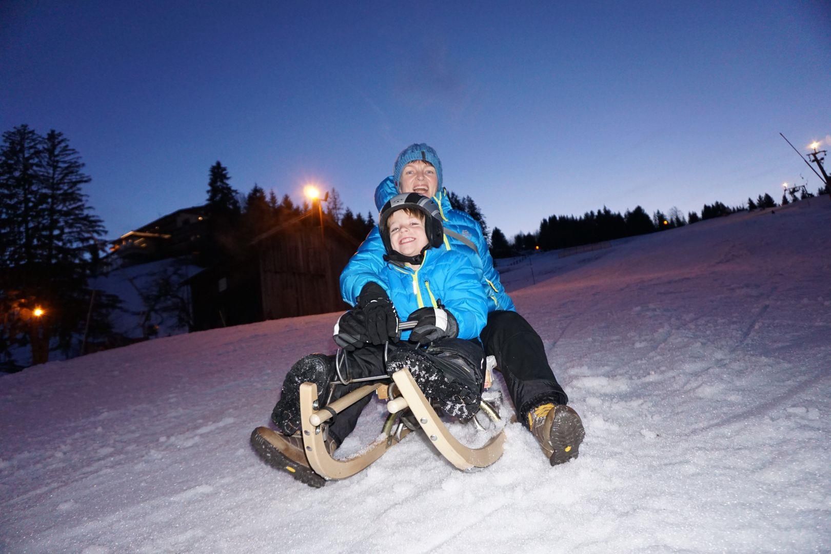 Rodeln am Abend am Stinesser-Lift, ein Erwachsener und ein Kind auf einem Schlitten im Schnee.