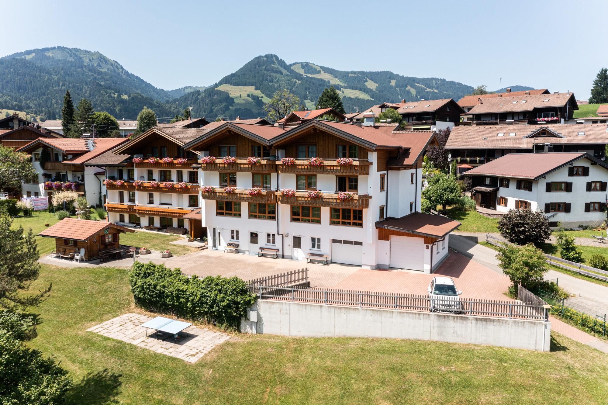 Mutterkindkurheim Marianne: Mehrstöckiges Gebäude mit Balkonen, Blumenkästen, umgeben von Häusern, grüner Wiese, Bergen im Hintergrund.