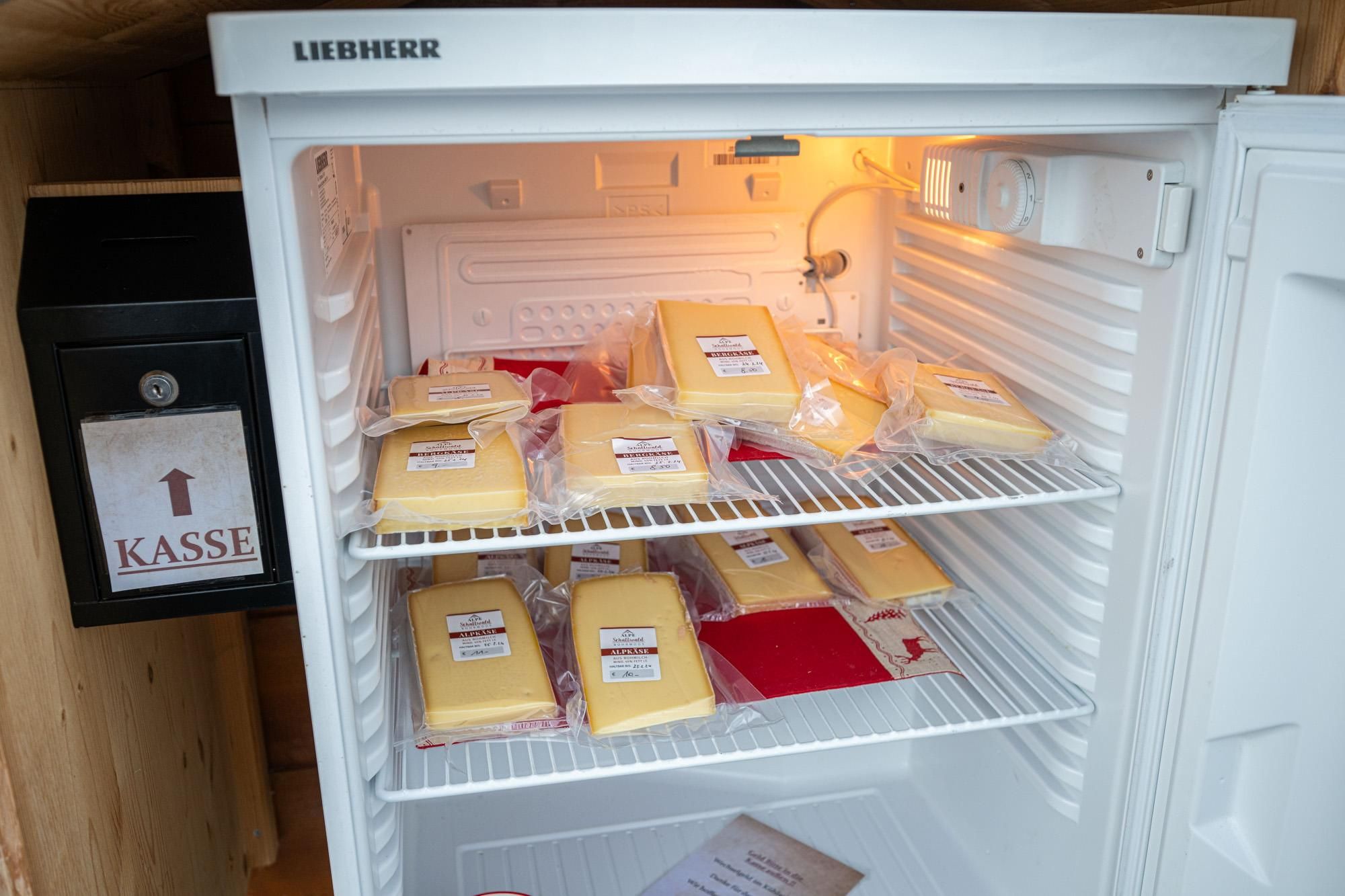Offener, weißer Kühlschrank gefüllt mit vakuumverpacktem Käse auf zwei Ebenen. Links daneben eine schwarze Kasse mit einem Pfeil nach oben und der Aufschrift "KASSE".