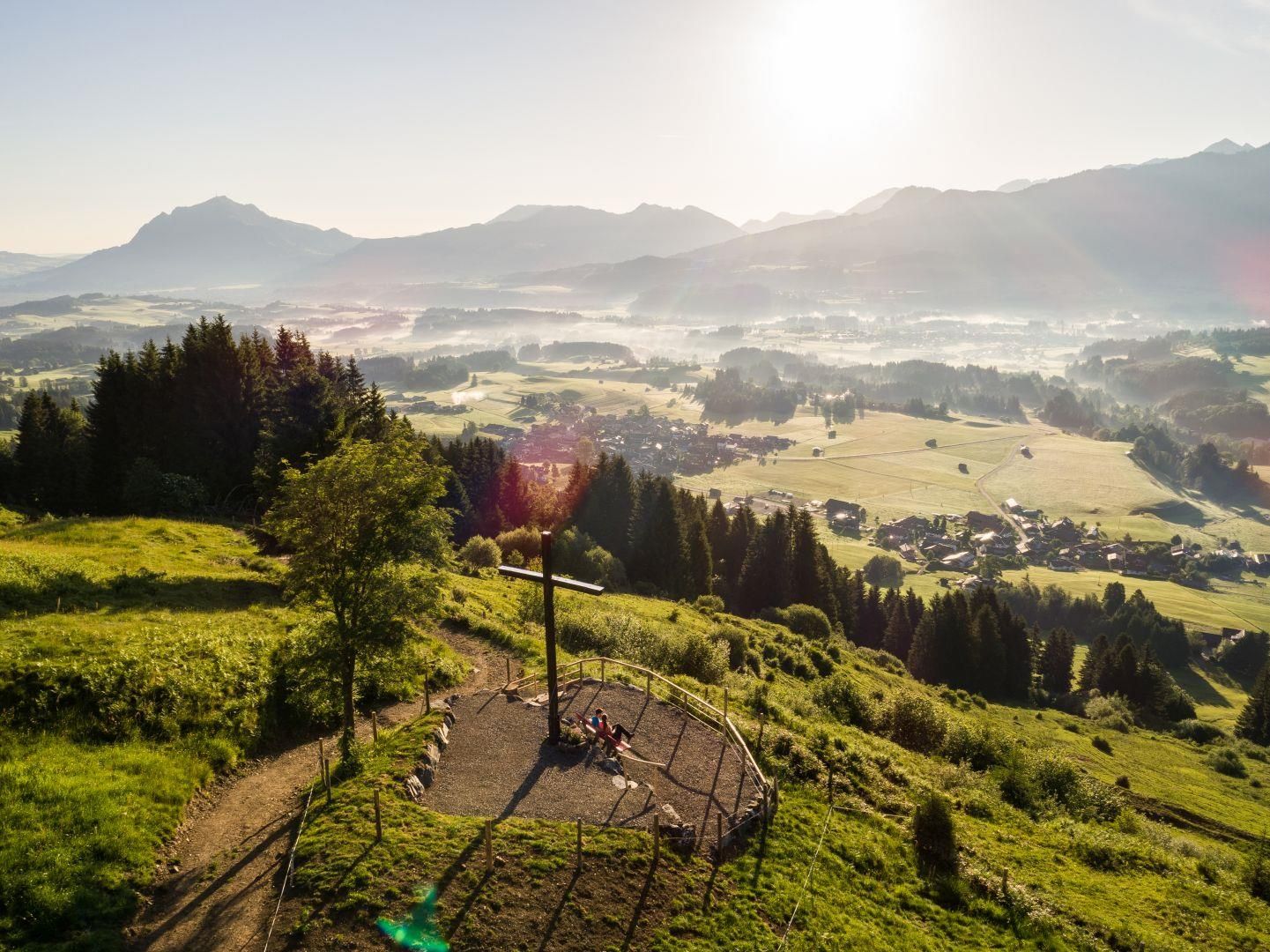 Hohes Holzkreuz auf einem Hügel. Umgeben von Wiesen und Wäldern. Im Hintergrund erheben sich majestätische Berge, teilweise in Nebel gehüllt.